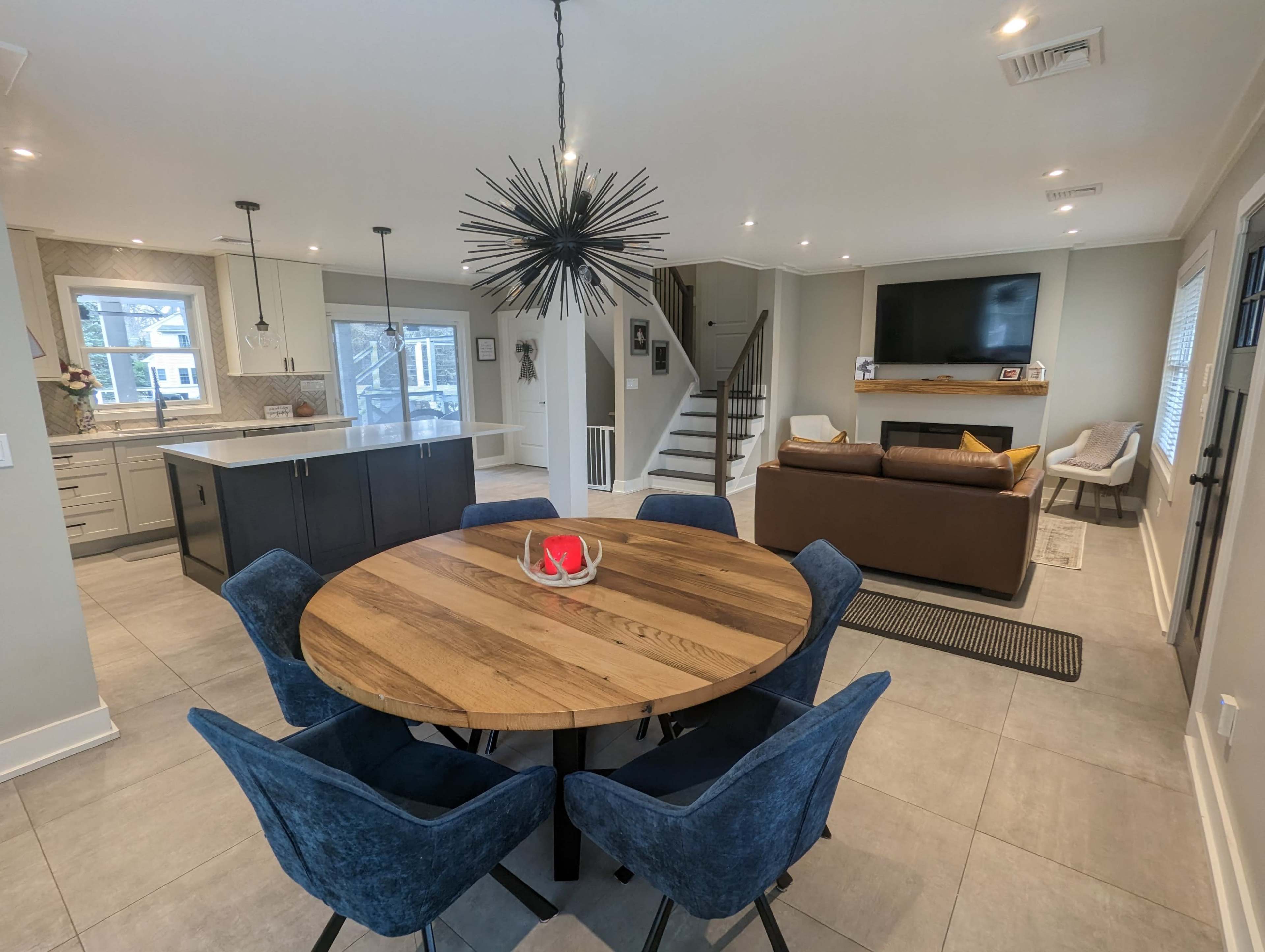 A modern dining area features a round wooden table surrounded by blue chairs, adjacent to a kitchen with a large island and stairs leading to an upper level.