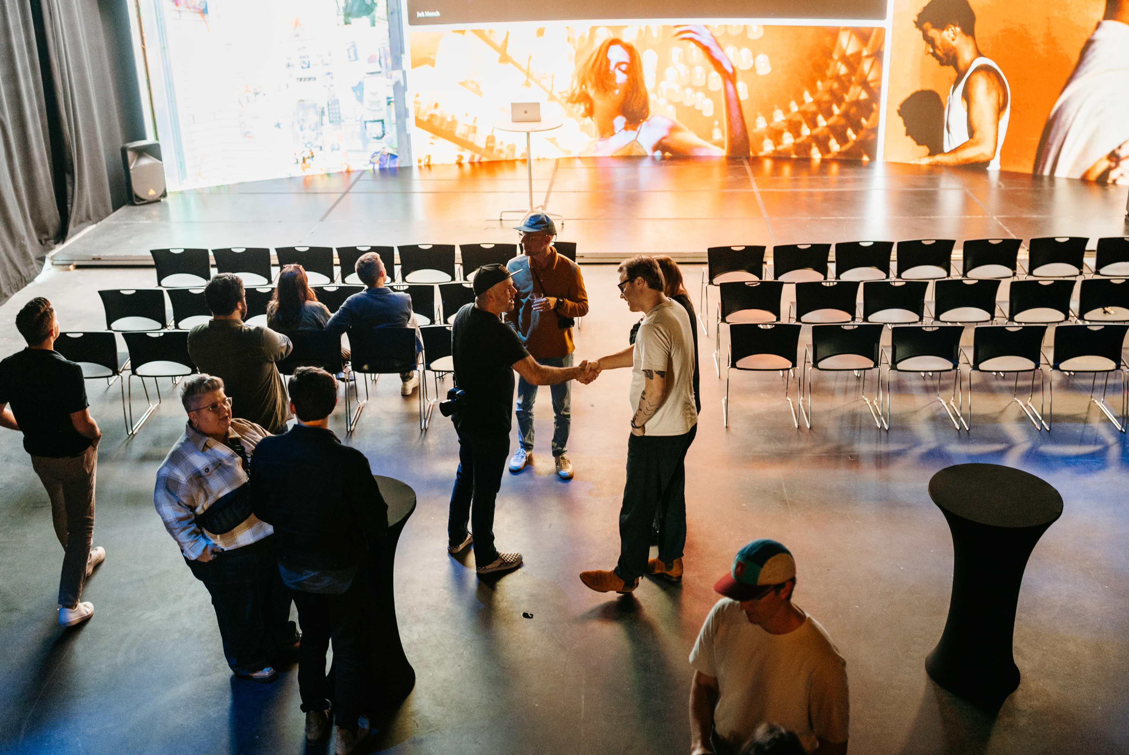 A group of people engages in conversation in a well-lit event space with empty chairs arranged in rows.