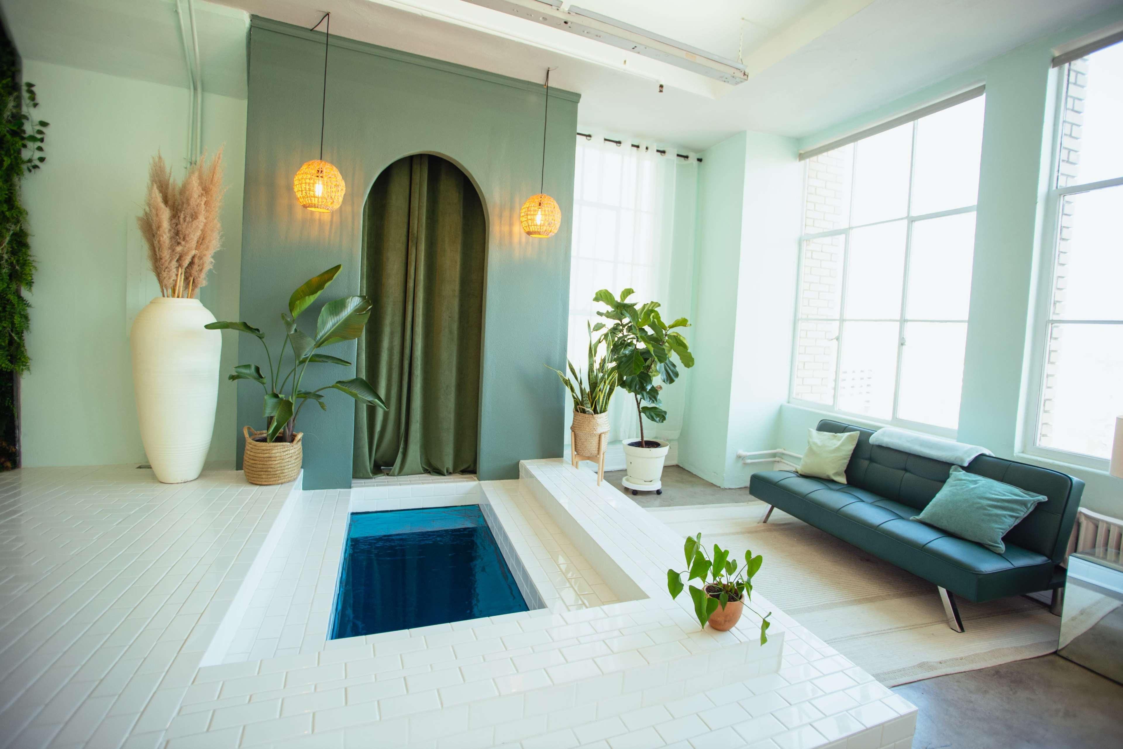 The image features a minimalist interior with a small pool, potted plants, and a green sofa against a backdrop of large windows and pastel walls.