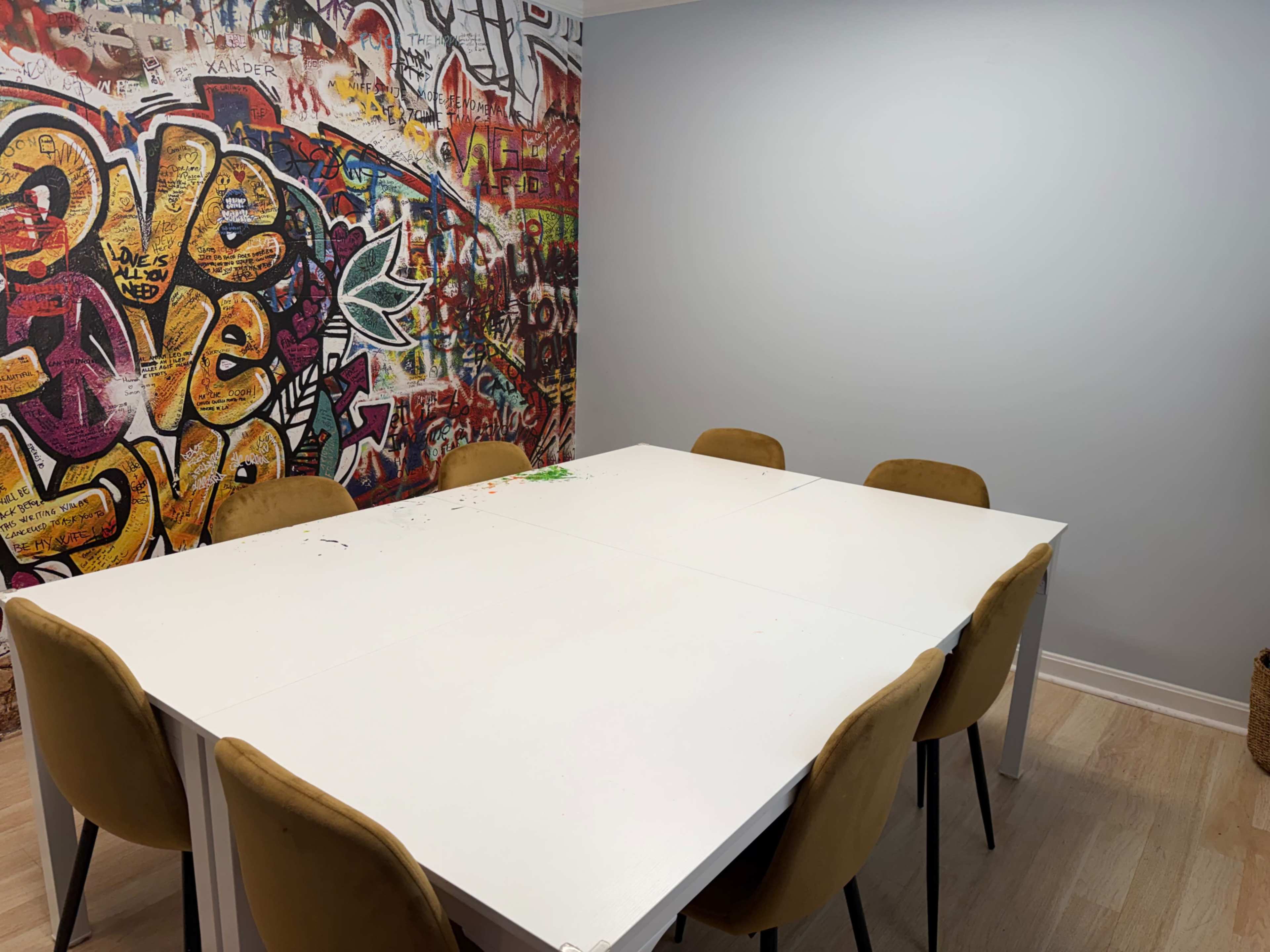 A white table with six brown chairs is positioned in front of a colorful graffiti-covered wall.