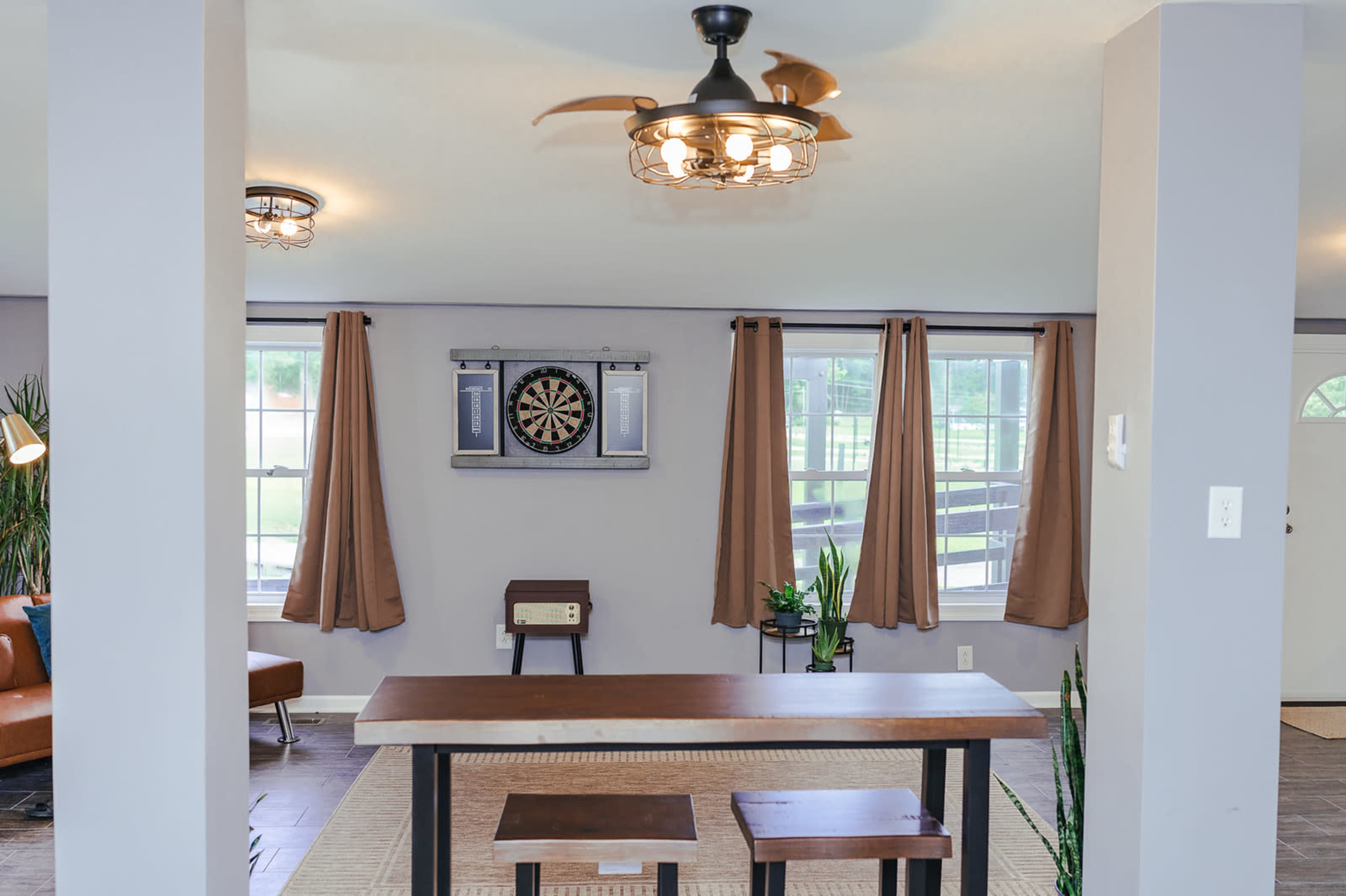 A spacious room with a wooden table and two stools in the center, a dartboard mounted on the wall, and large windows with curtain drapes.