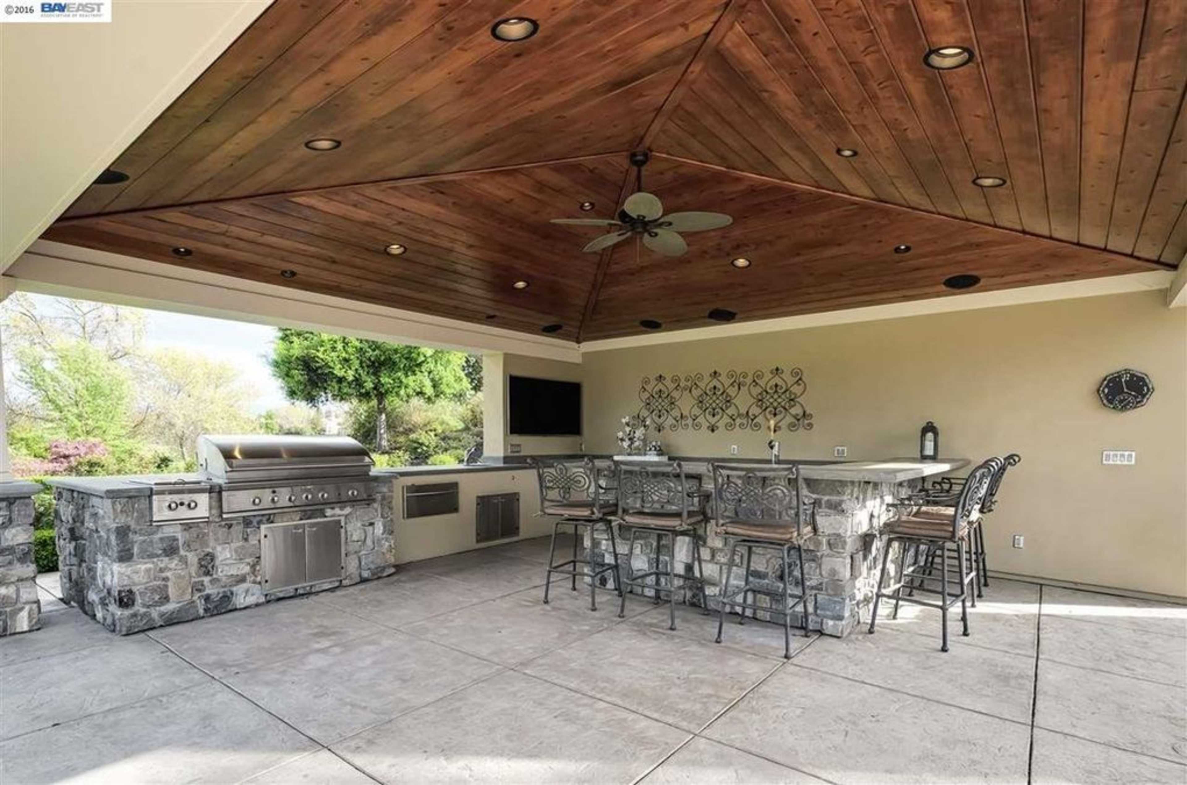 The image shows an outdoor kitchen and dining area with a stone grill, bar seating, and a wooden ceiling.