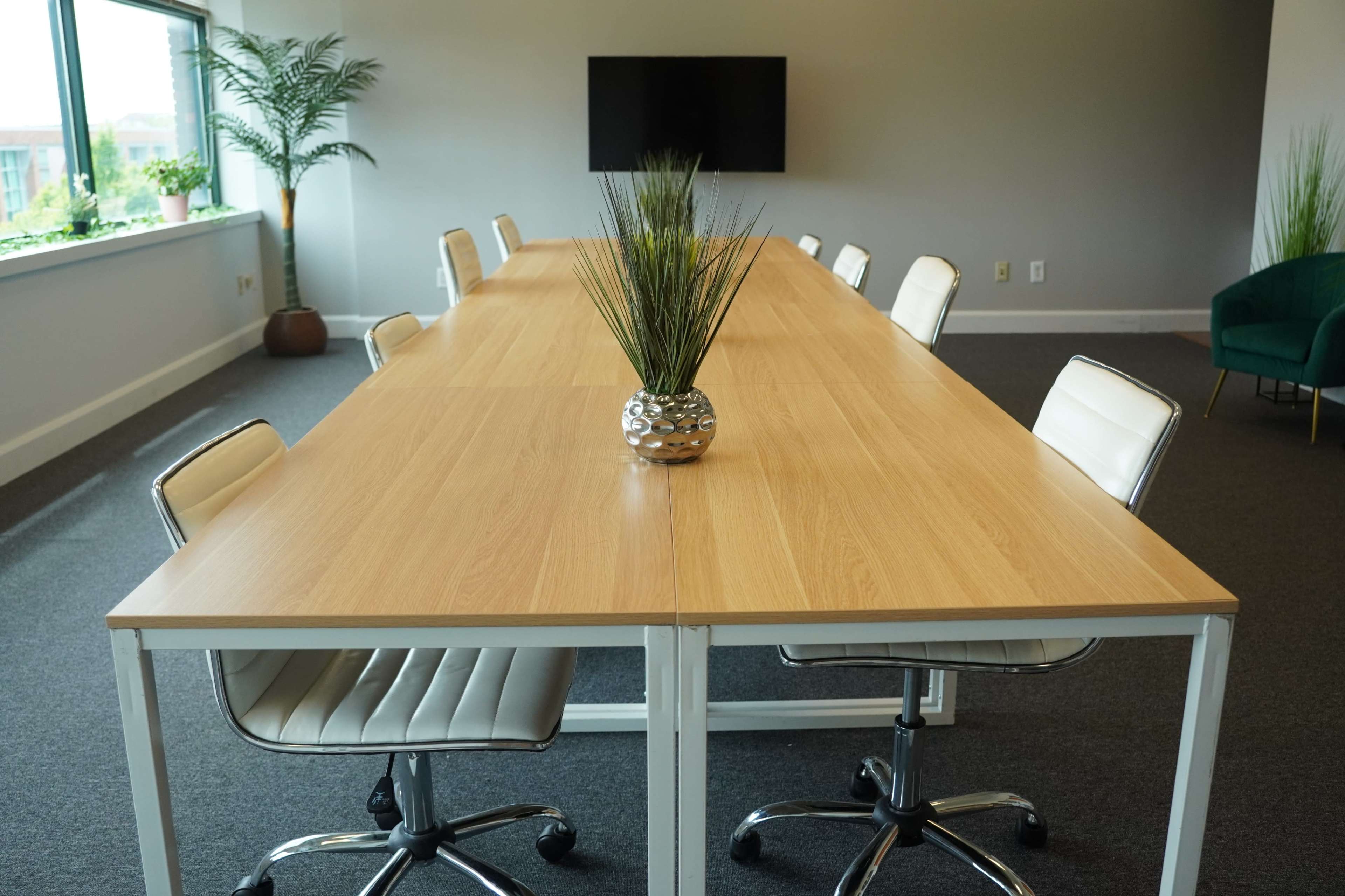 A long wooden conference table with white chairs is set in a well-lit meeting room featuring a large window and a television screen on the wall.
