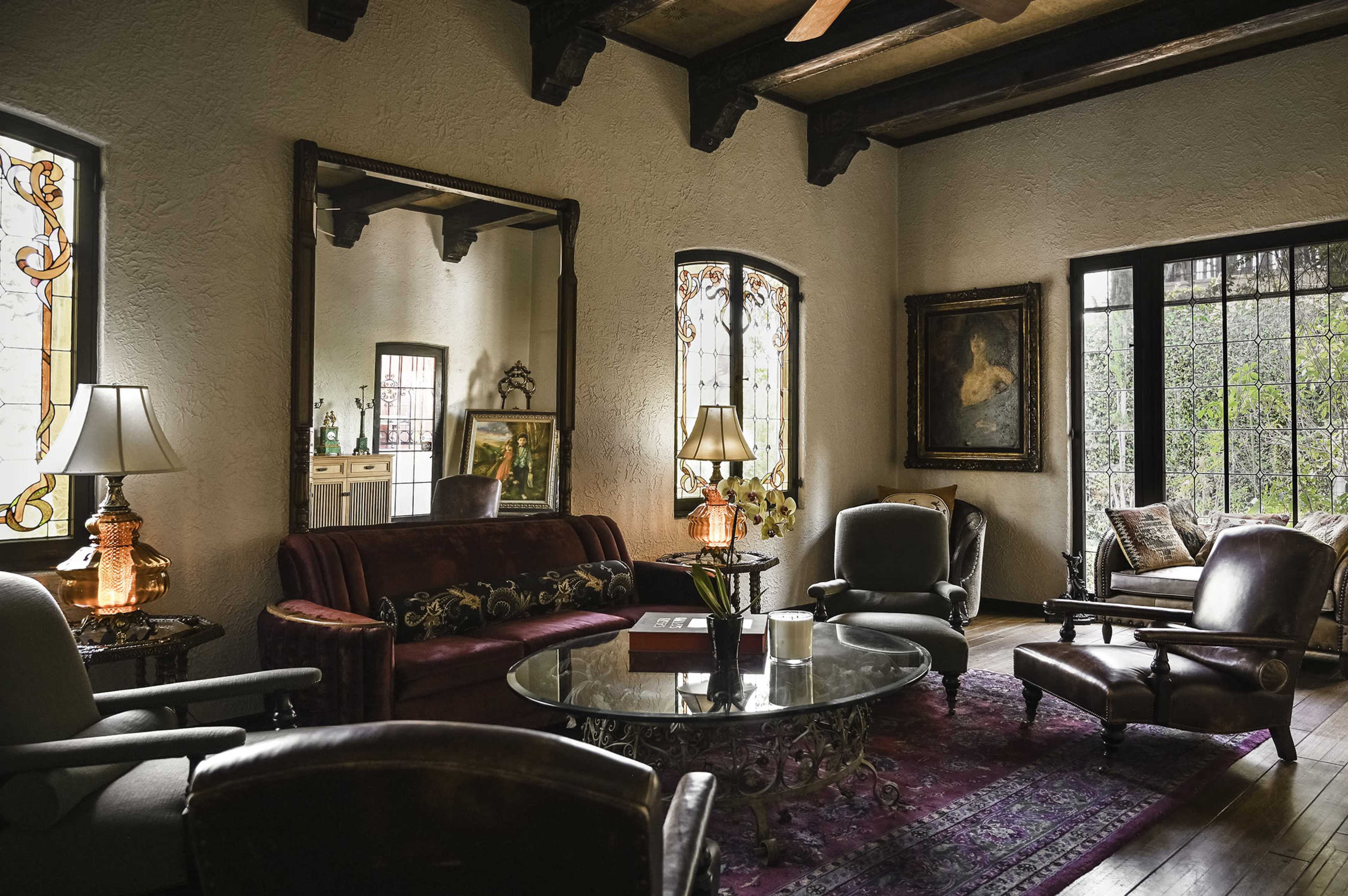 The living room features a mix of vintage furniture, including a maroon couch, several armchairs, and a glass coffee table surrounded by large windows and ornate wall decor.