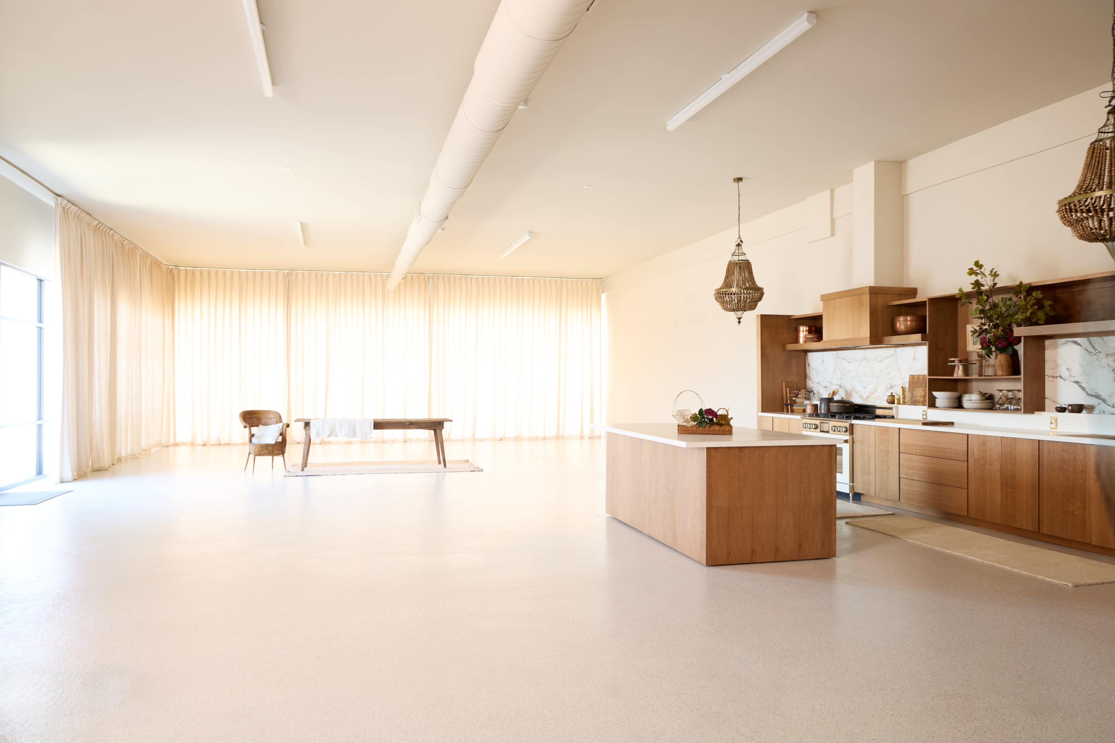 The image shows a spacious, modern kitchen with wooden cabinetry and countertops, complemented by a large table and abundant natural light from curtains hanging on the windows.