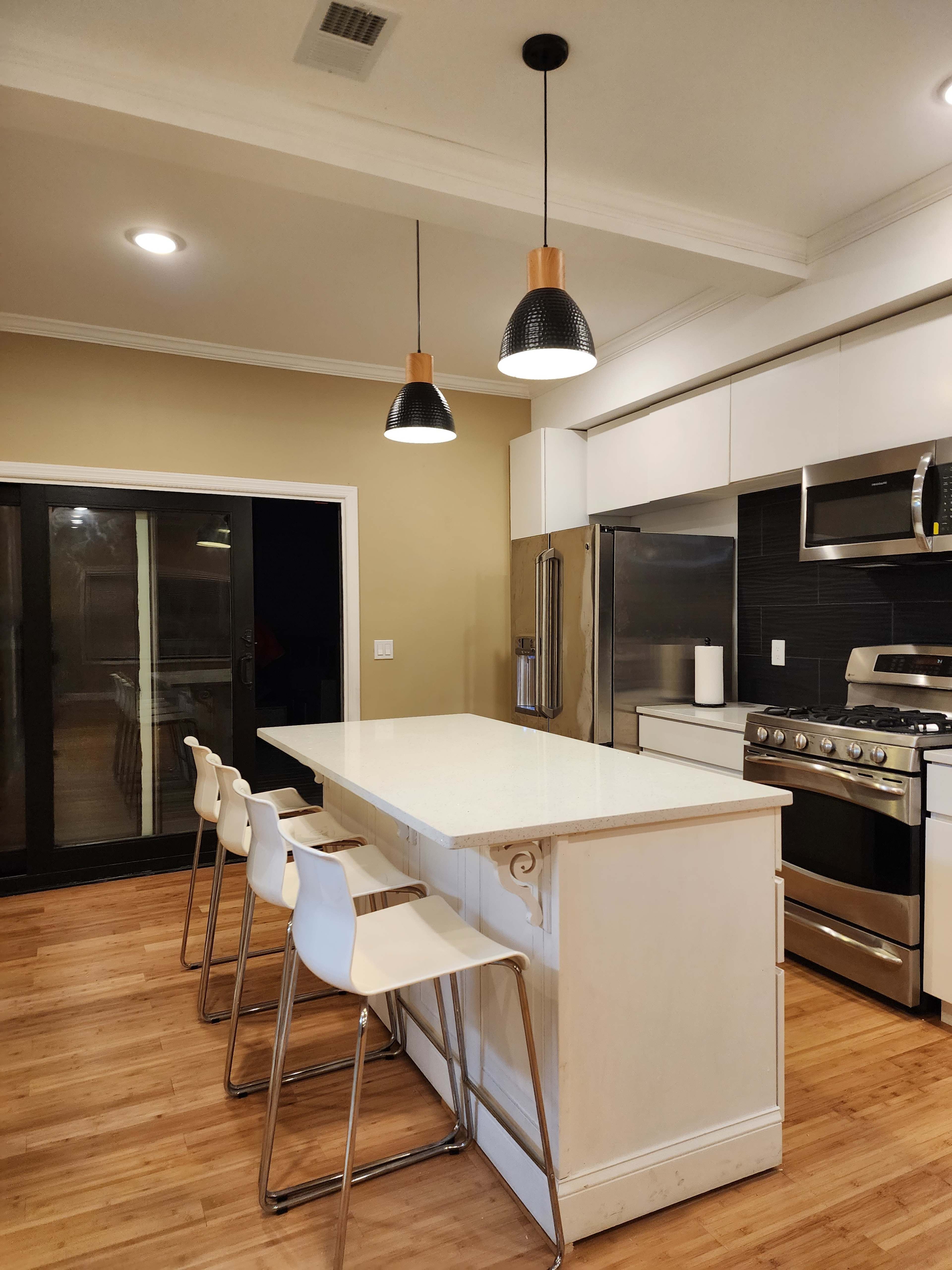 A modern kitchen features a large island with four white stools, stainless steel appliances, and three pendant lights overhead.
