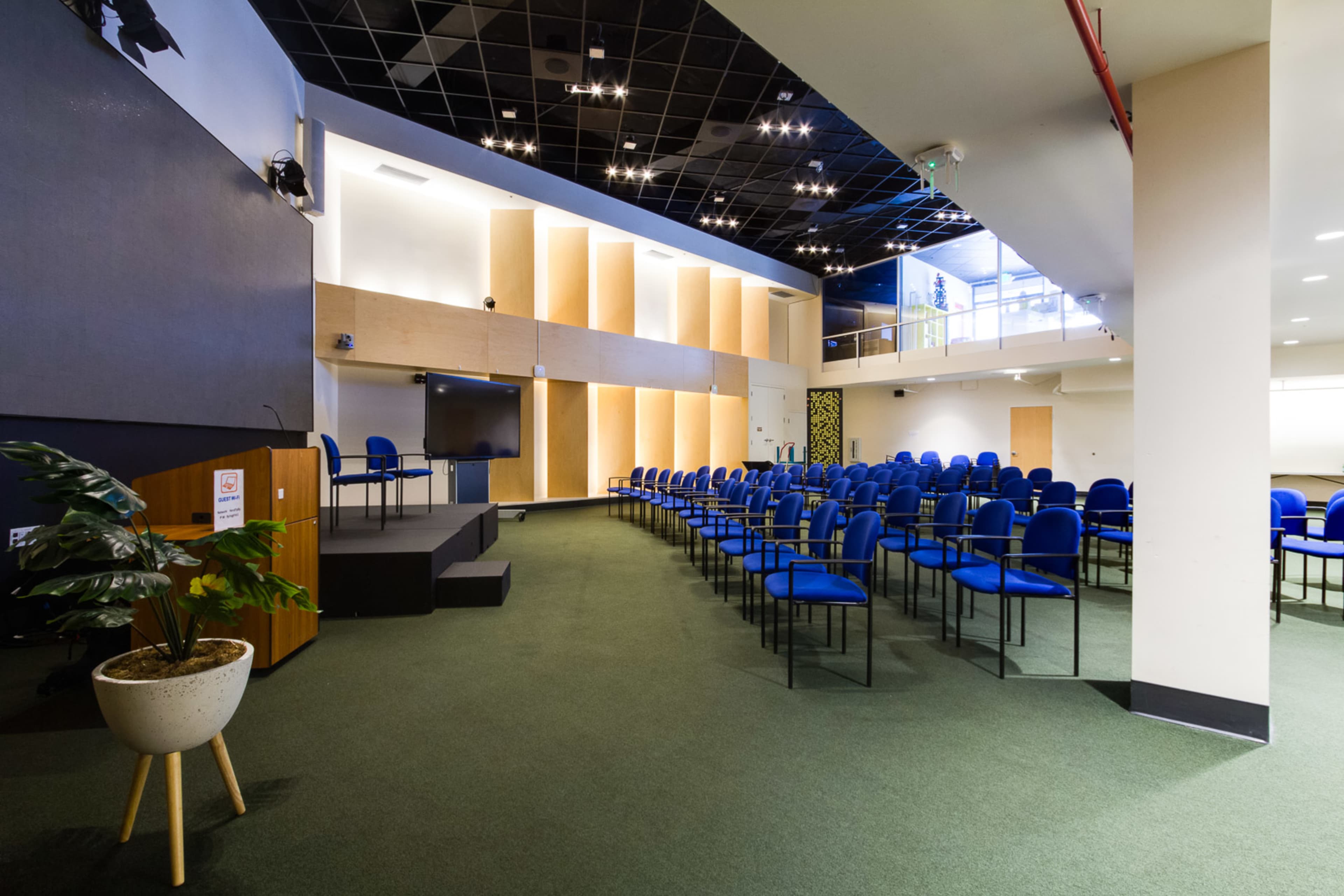 A spacious conference room with rows of blue chairs facing a podium, a screen, and a staircase leading to an upper level.