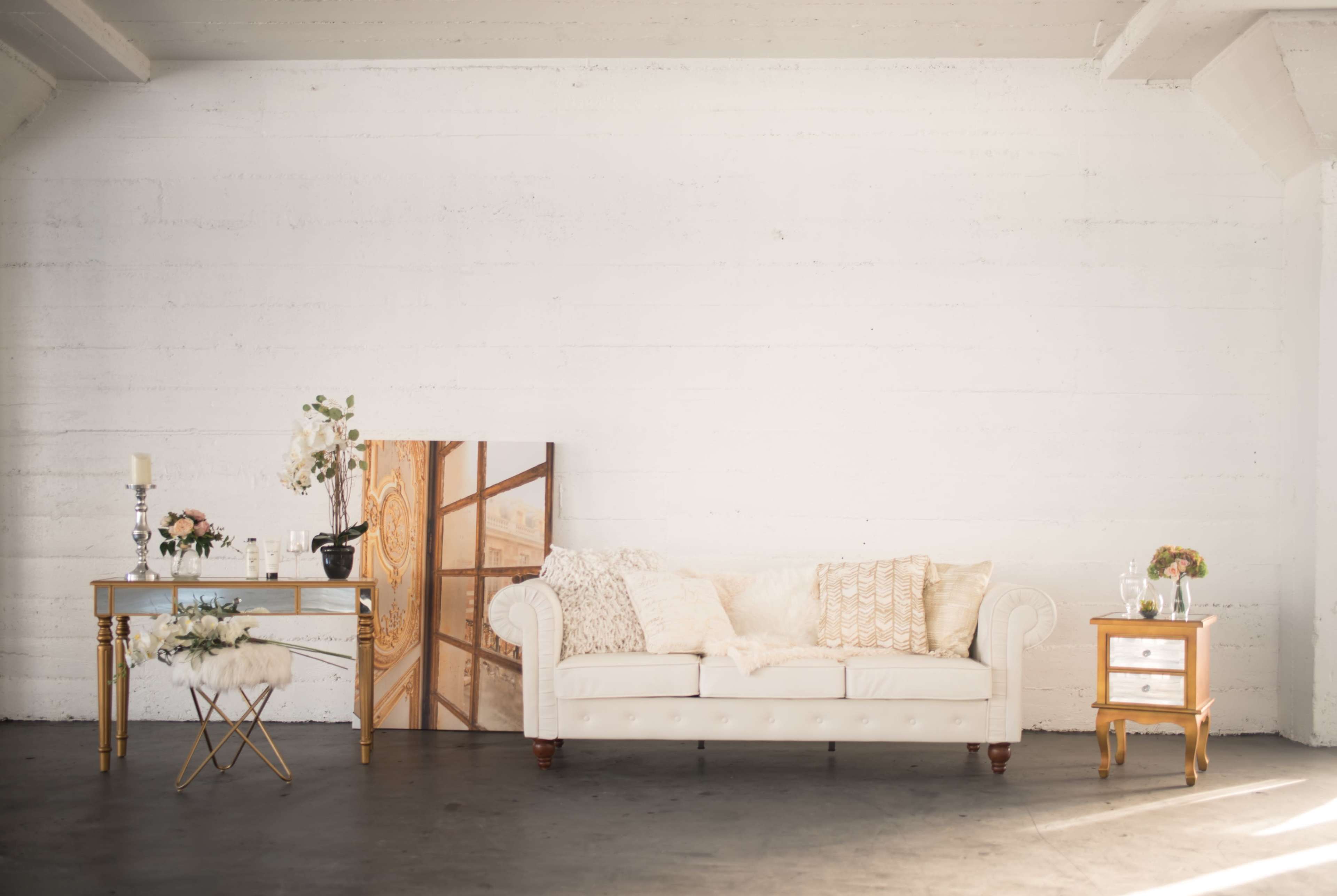 The image shows a light-colored couch with decorative pillows, positioned next to a small table and a bedside table in a minimalist interior space.