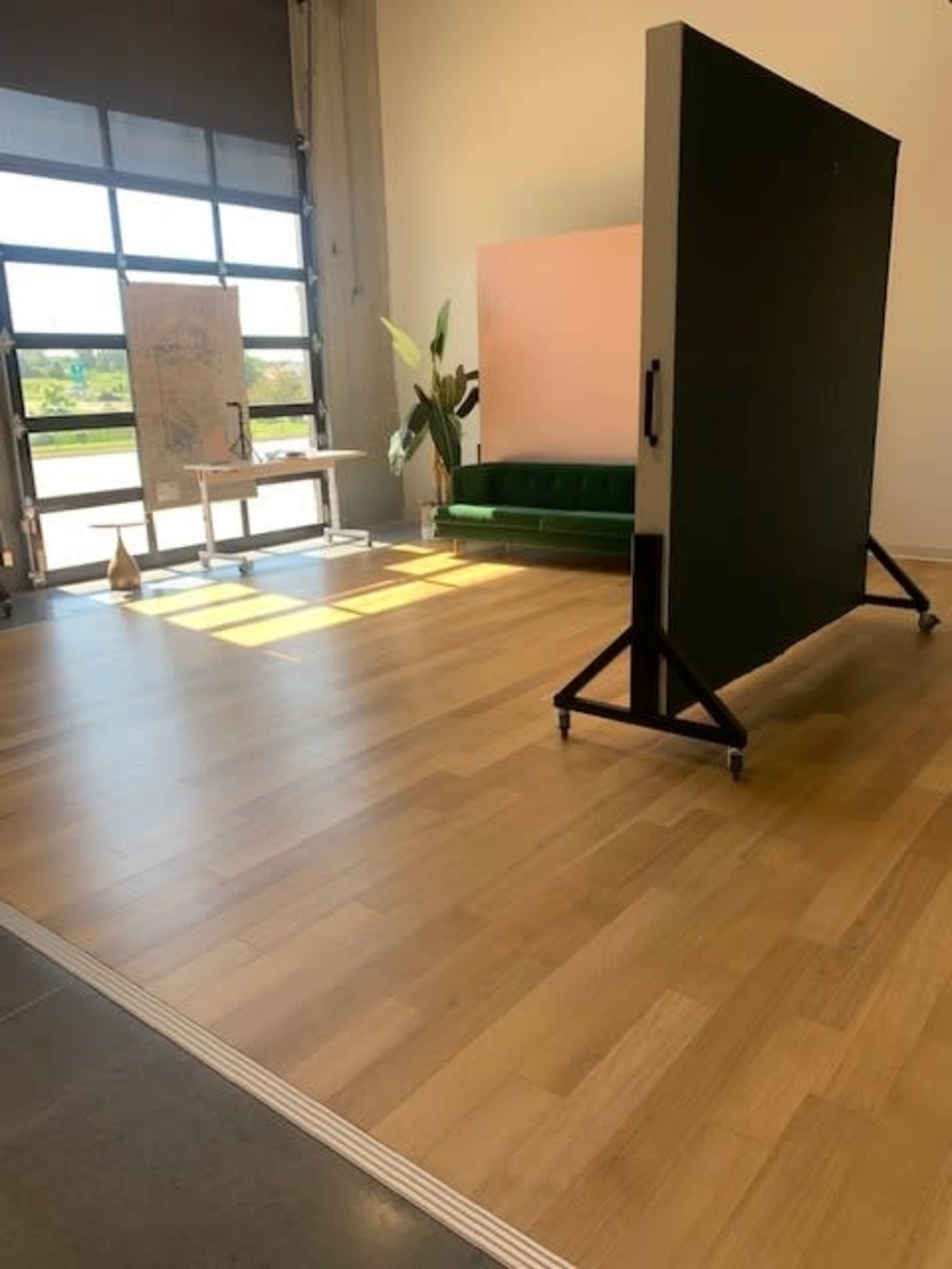A spacious interior with a green sofa, a large movable black panel, and a light wooden floor, illuminated by natural light from large windows.