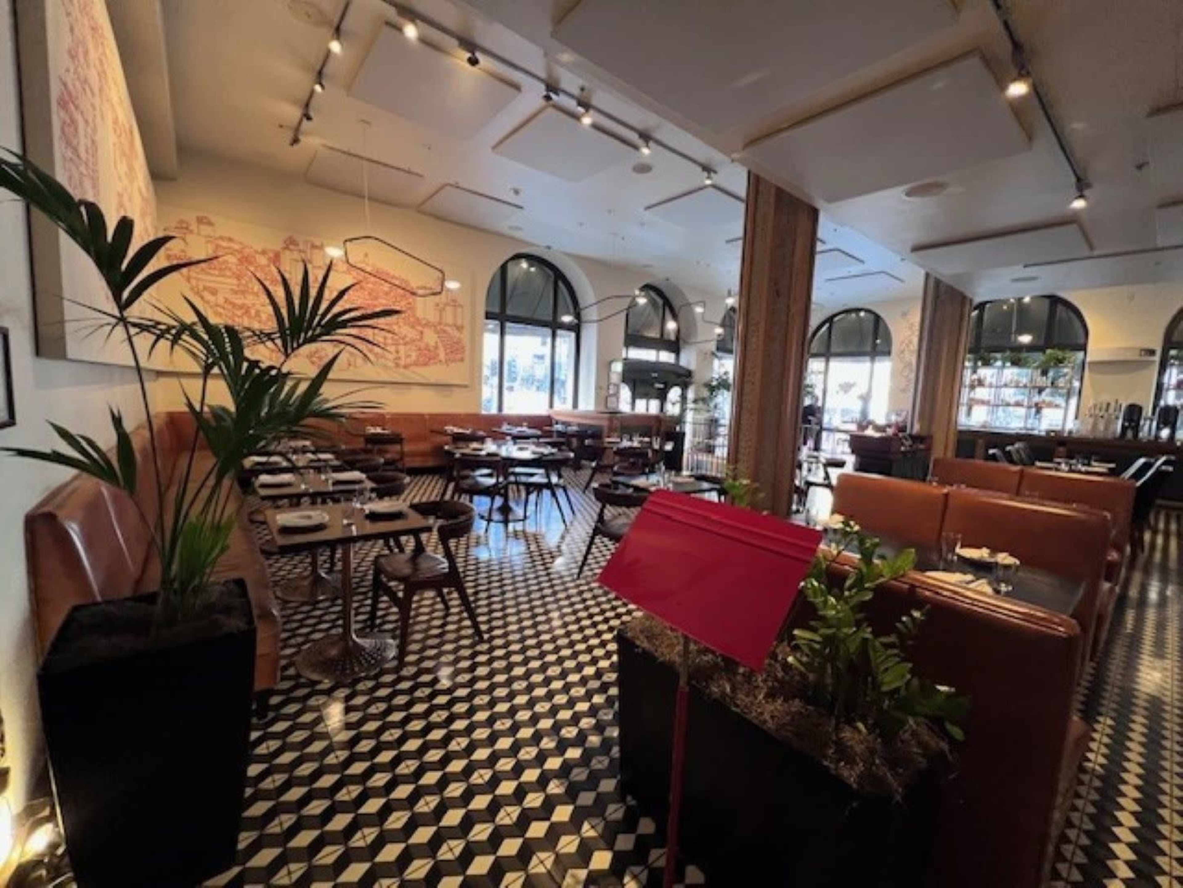 The image shows the interior of a restaurant featuring tiled floors, wooden tables, and large windows with decorative plants.
