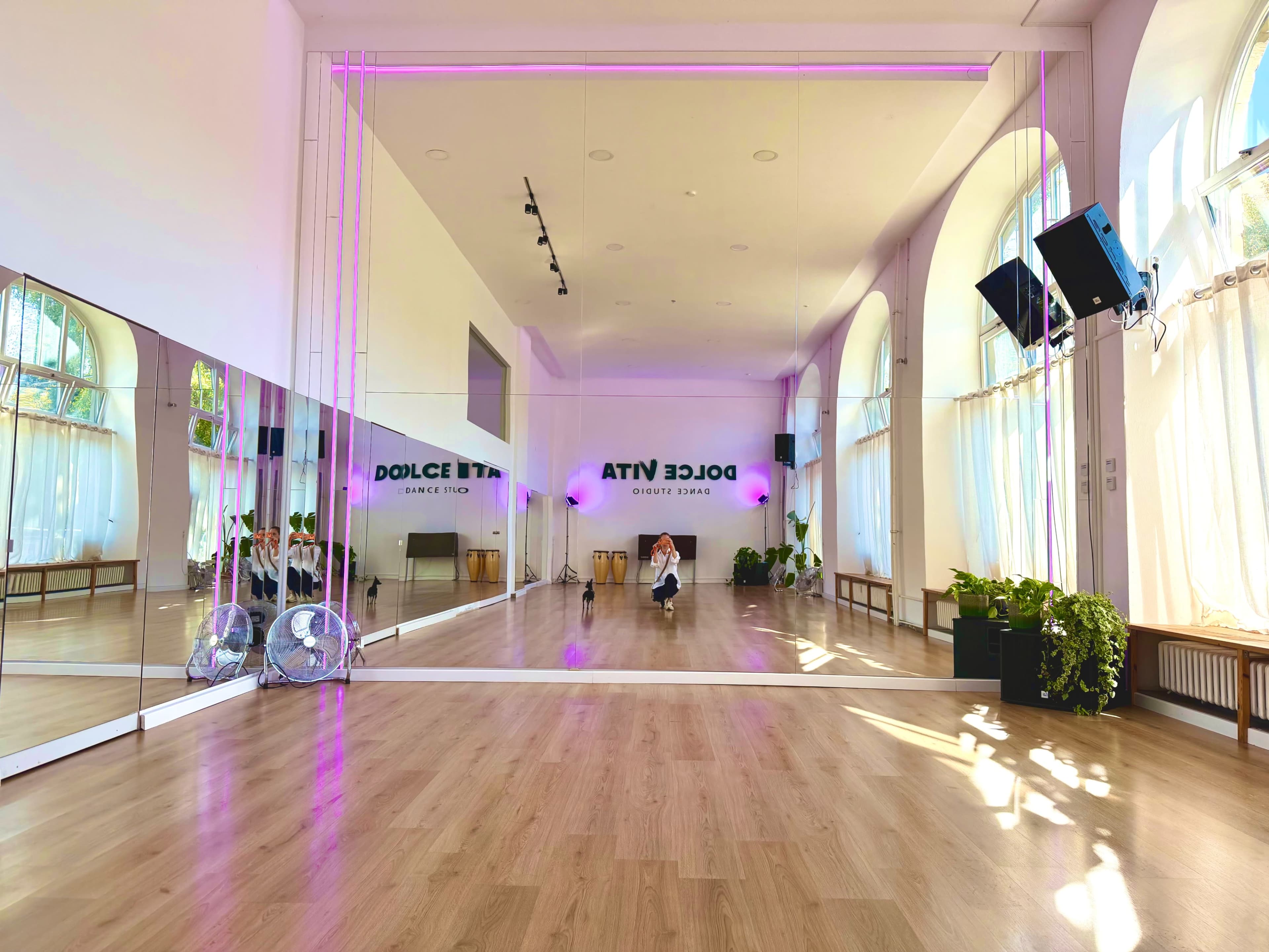 The image shows a spacious dance studio with large mirrors, wooden flooring, and purple lighting.