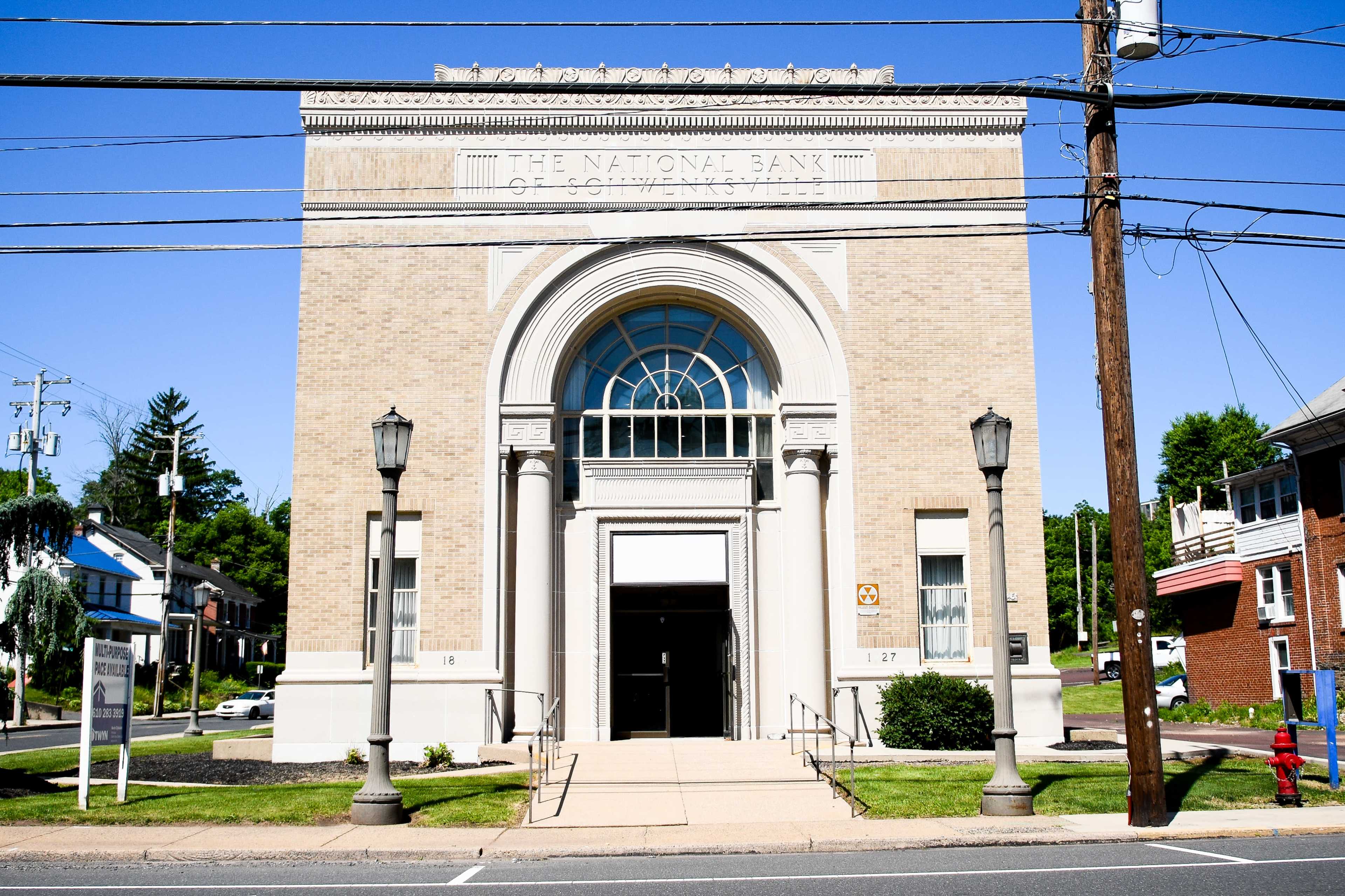 The image shows the exterior of a historic bank building featuring a large arched entrance and columns, with streetlamps and utility poles in the foreground.