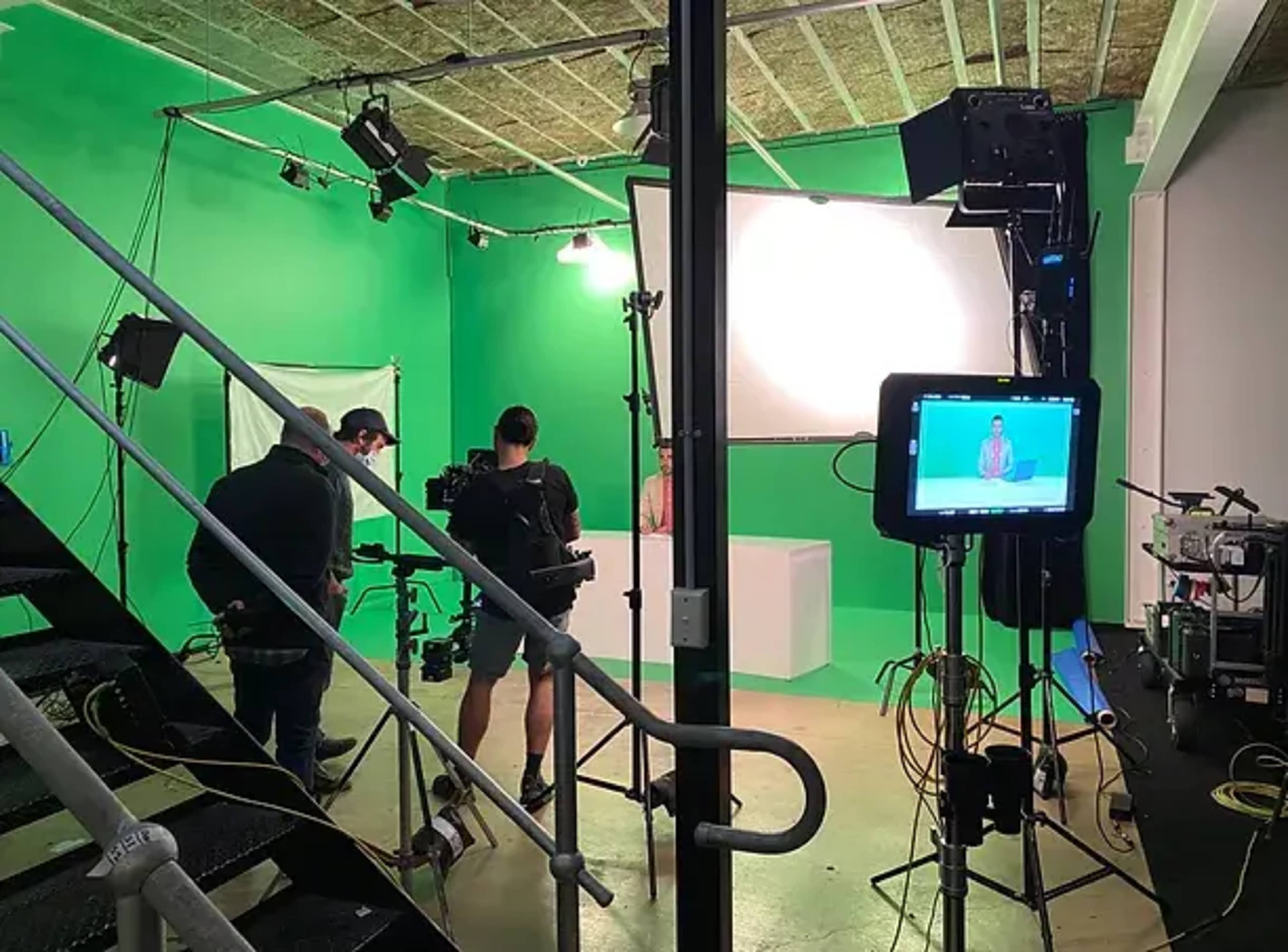 A filming crew works in a green-screen studio, with bright lights and cameras positioned around a central white podium.
