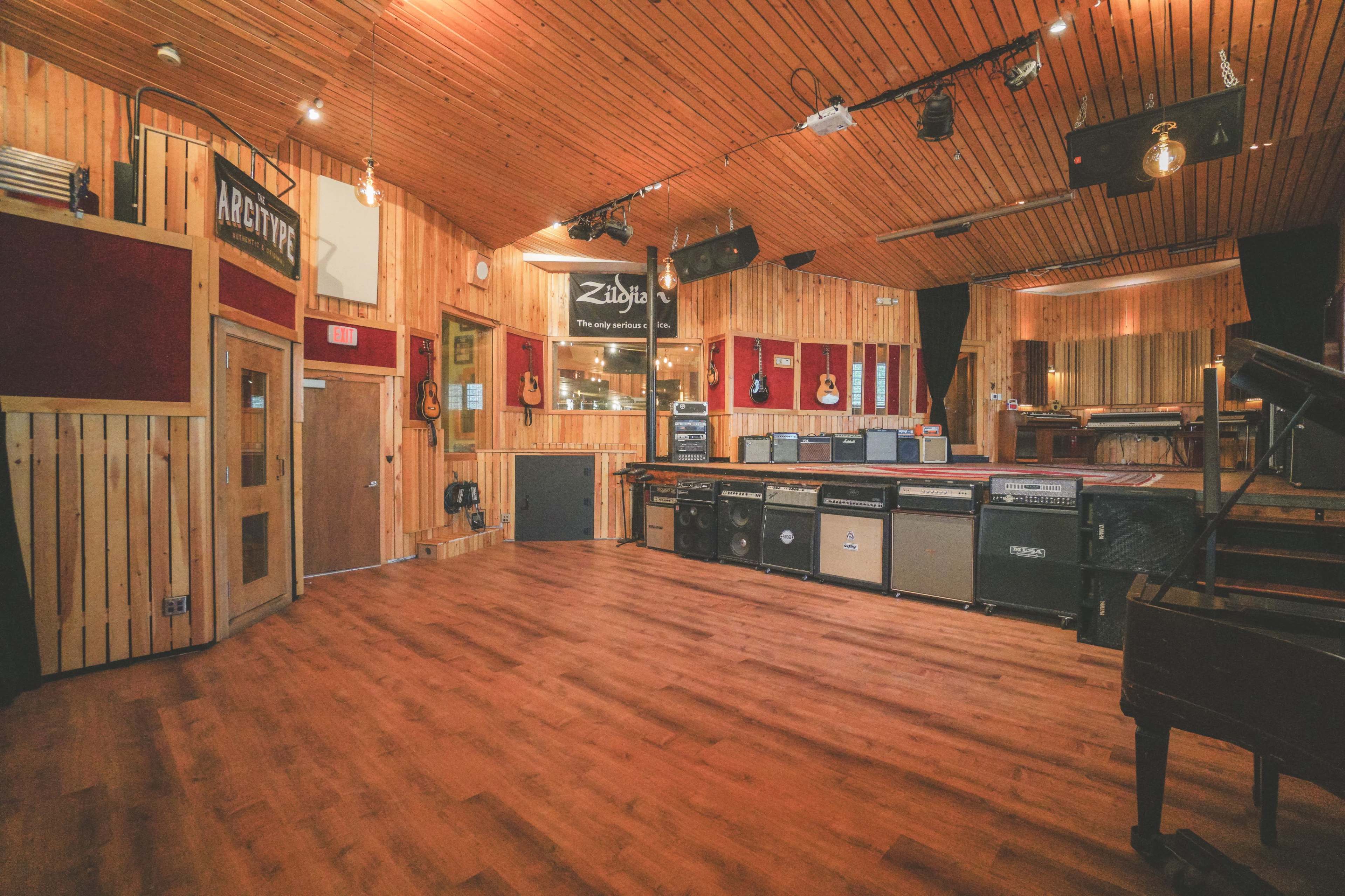 The image shows a spacious wooden-floored recording studio with acoustic panels, amplifiers lined against the wall, and a piano in the corner.