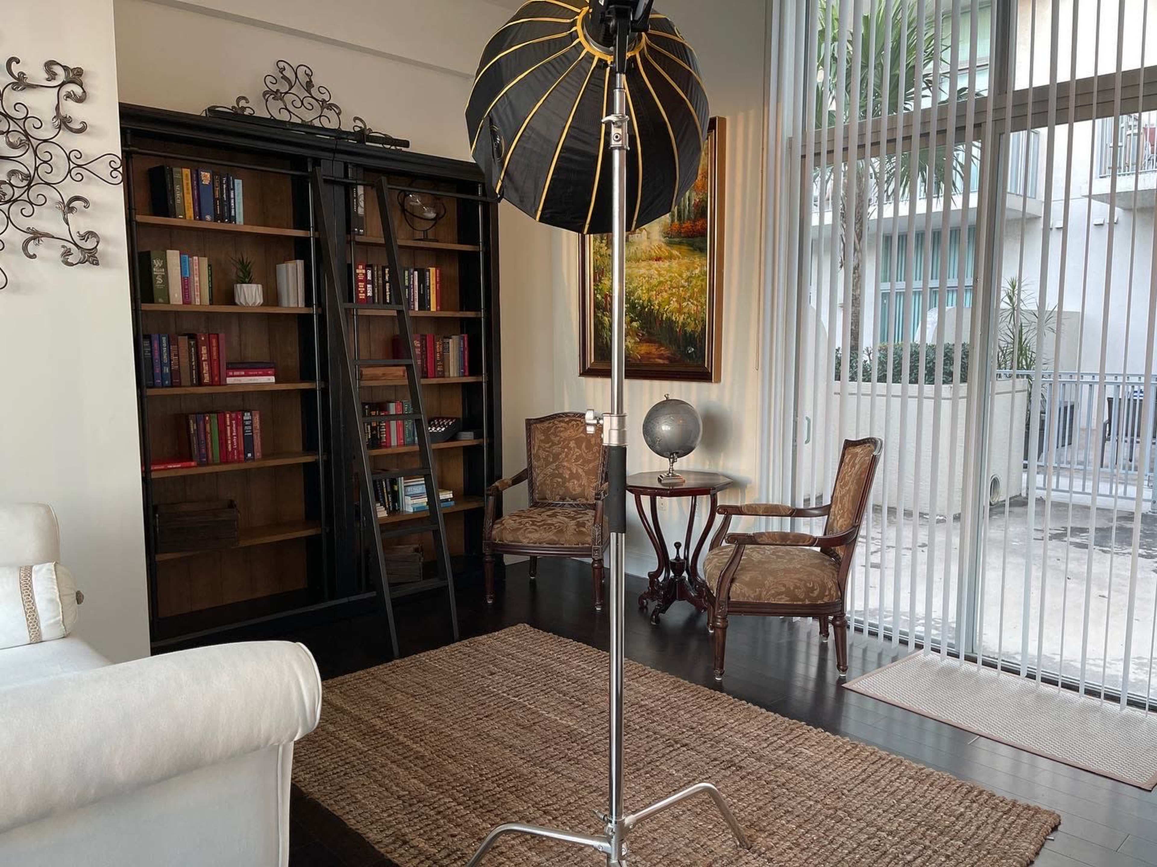 The image shows a well-lit living space featuring a black bookshelf filled with books, two ornate chairs, a small round table with a globe, and large sliding glass doors leading to an outdoor area.