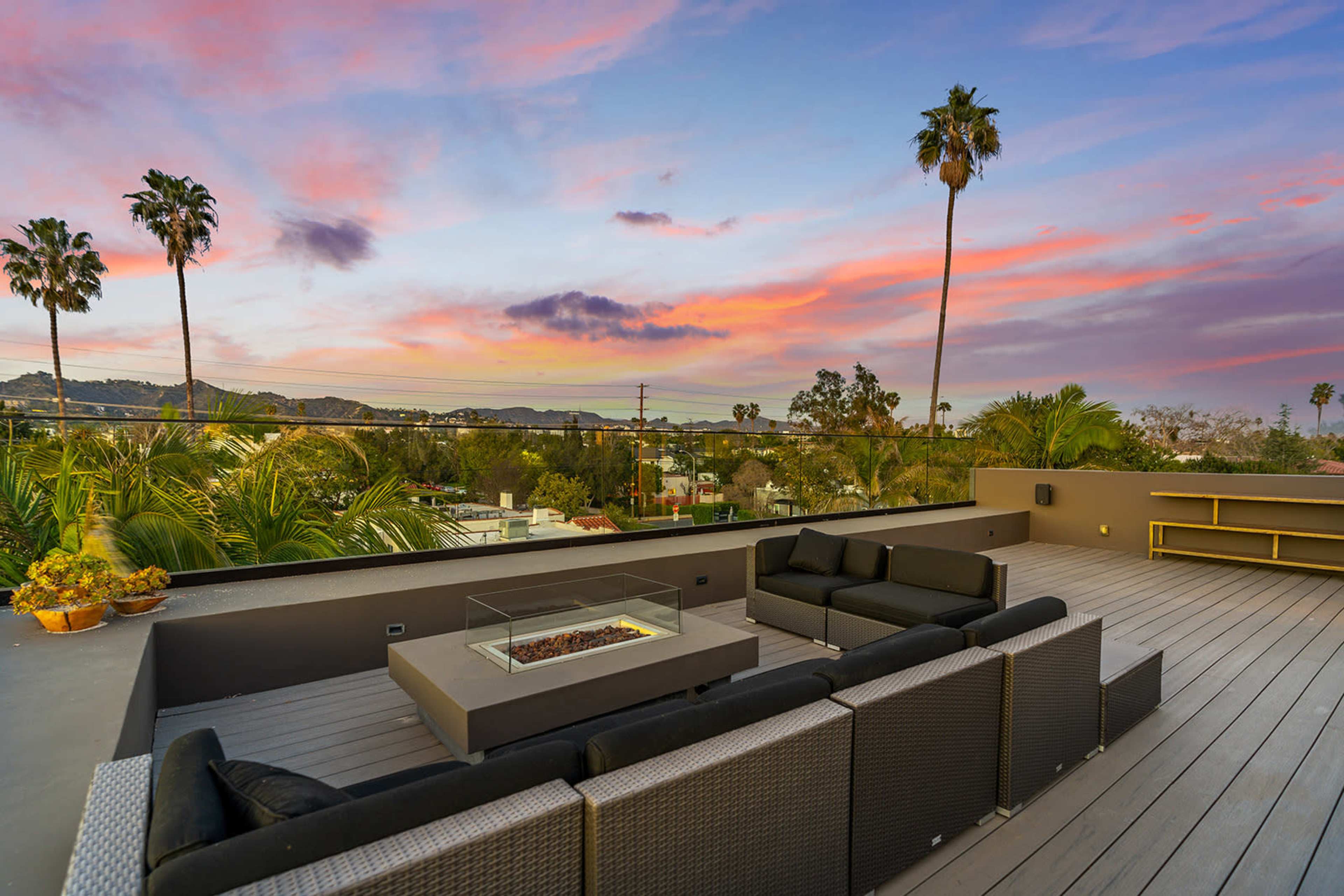 A spacious rooftop patio features a seating area around a fire pit, with palm trees and a colorful sunset in the background.