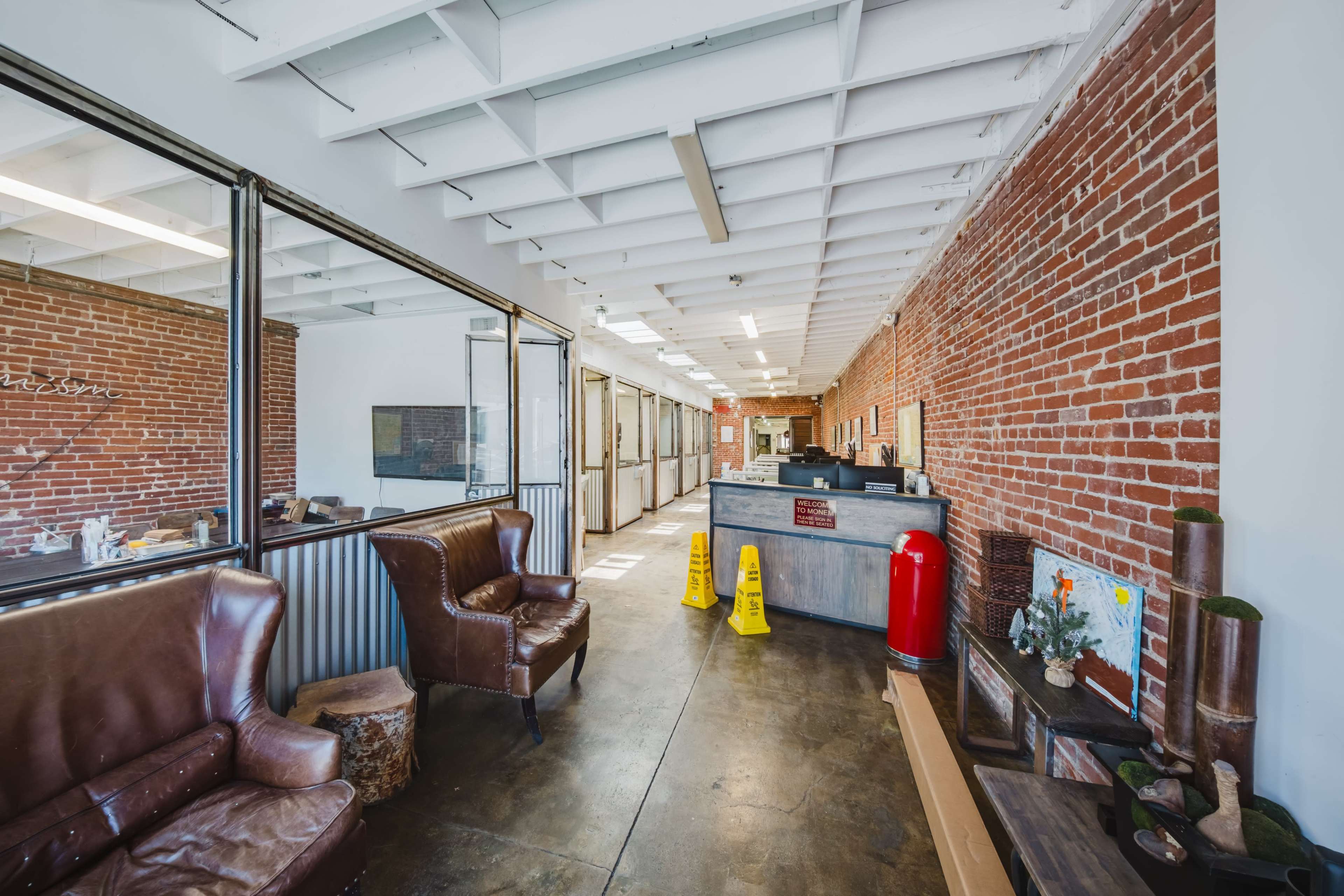The image shows a modern office hallway with exposed brick walls, a reception area, and seating arrangements including leather chairs and a wooden table.