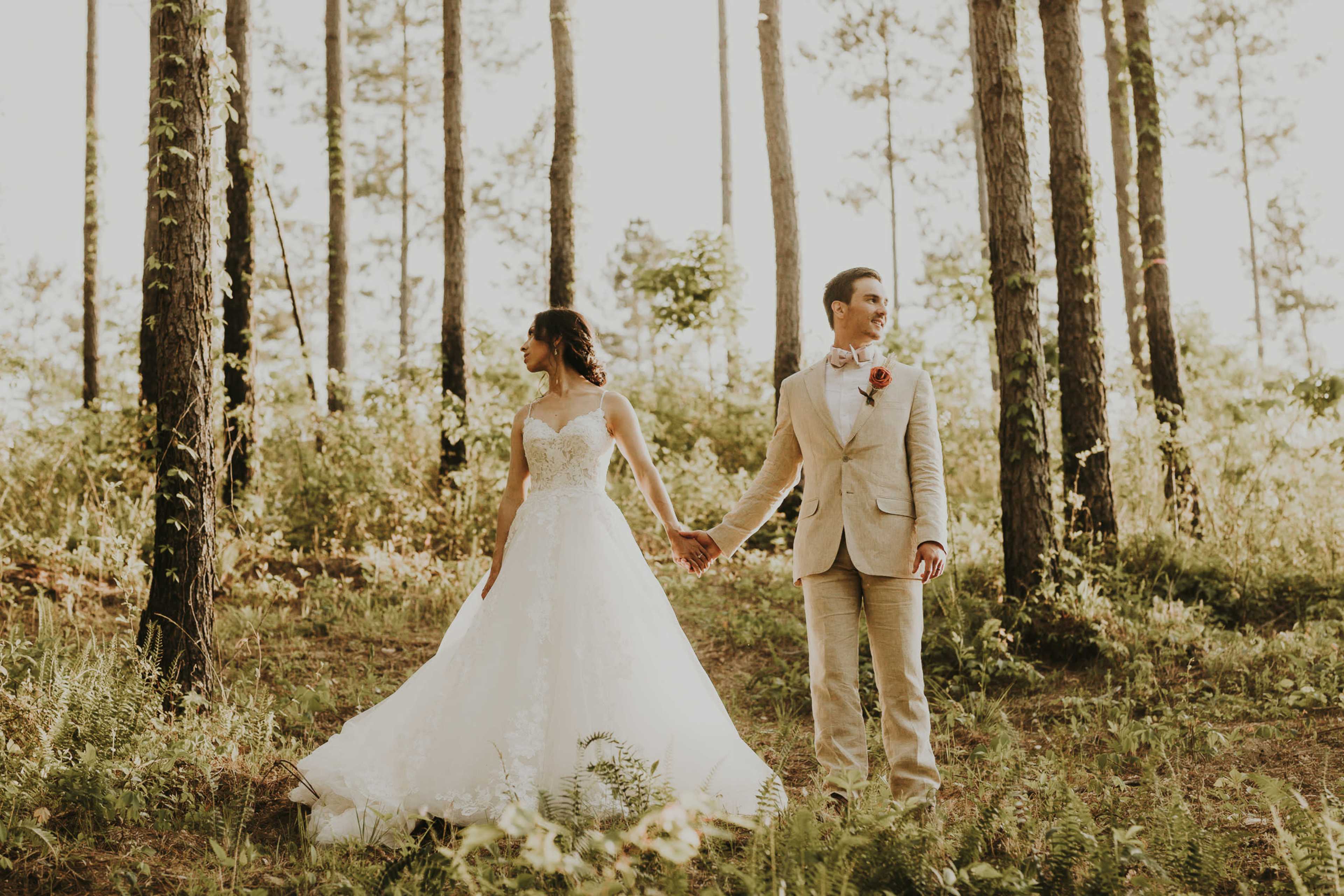 A bride and groom hold hands in a wooded area, surrounded by tall pine trees.