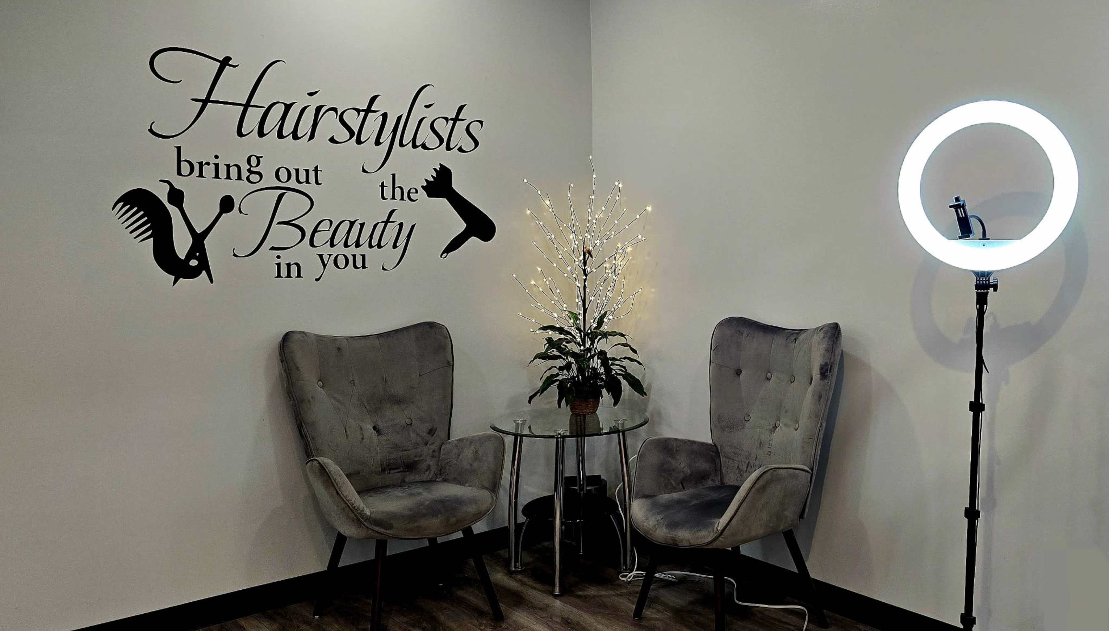 The image depicts a cozy salon corner featuring two upholstered chairs, a small glass table with a plant, and a light ring next to a decorative wall with the phrase "Hairstylists bring out the Beauty in you."