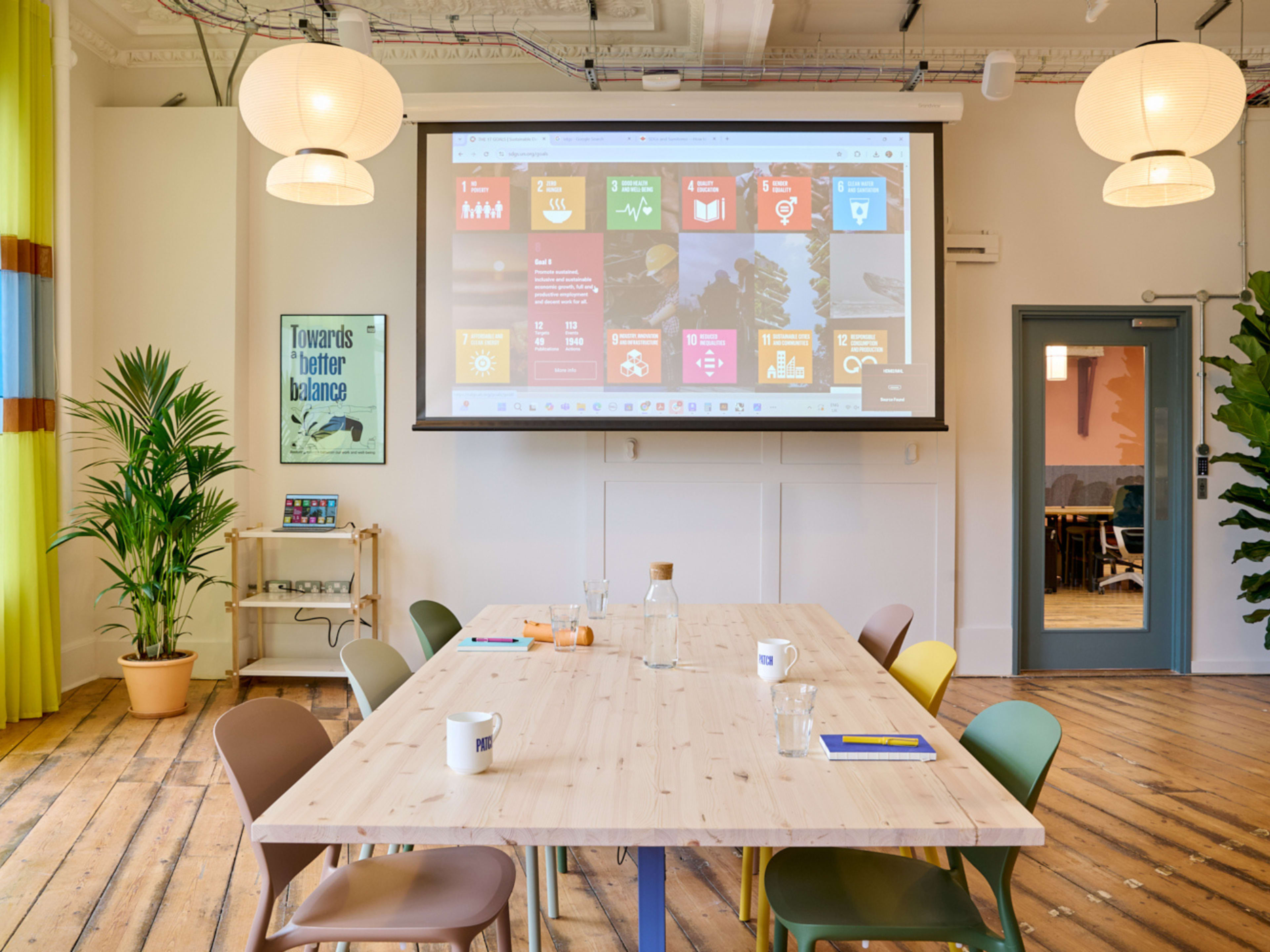 A large wooden table surrounded by colored chairs is set in a bright room featuring plants, a projector screen displaying colorful slides, and ambient lighting.