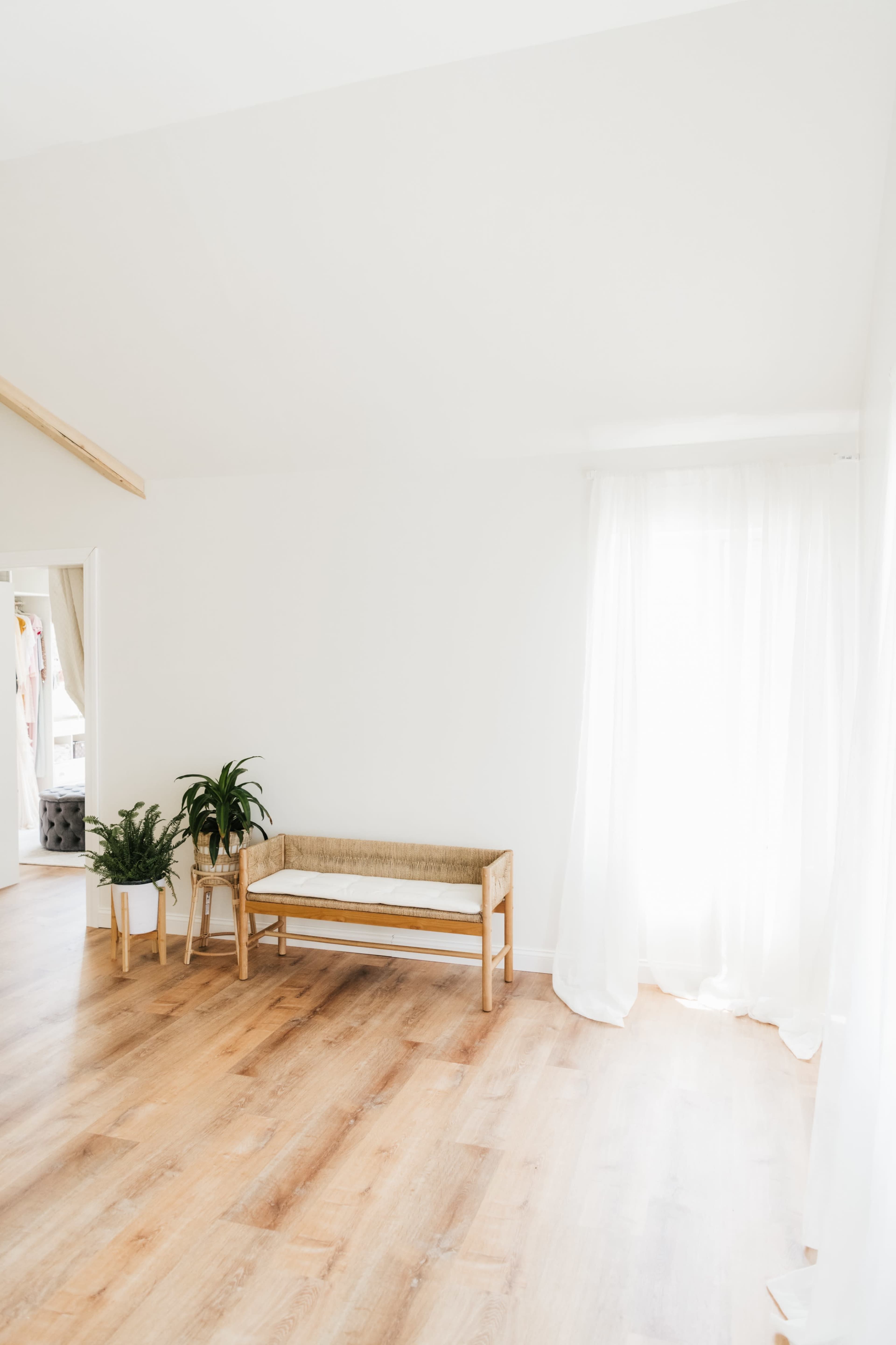 A minimalist room features a wooden bench and potted plants beside a large window with sheer curtains.