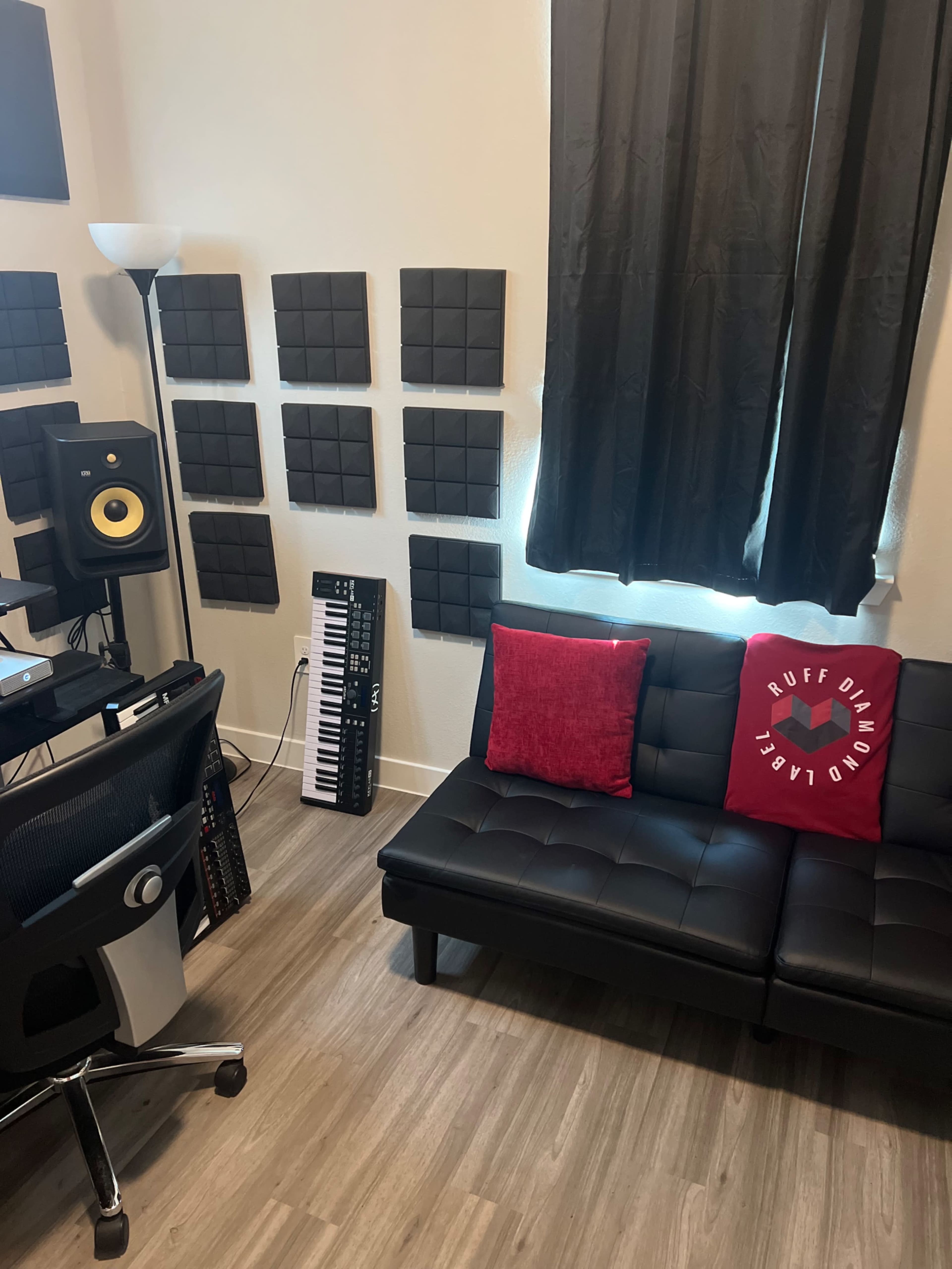 A music production studio featuring a black couch, a keyboard, acoustic panels on the walls, and a workstation with a lamp.