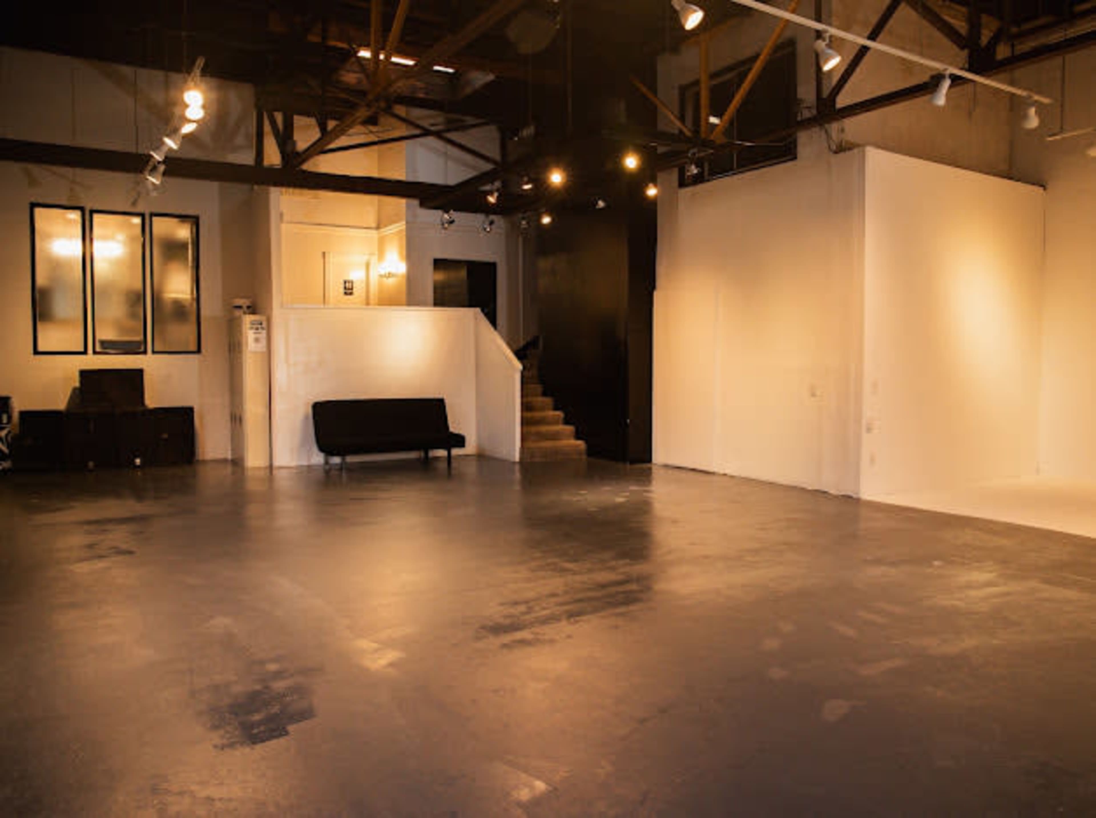 The image shows a spacious, empty gallery with polished concrete floors, high ceilings, and minimalistic decor, including a black sofa and a staircase.