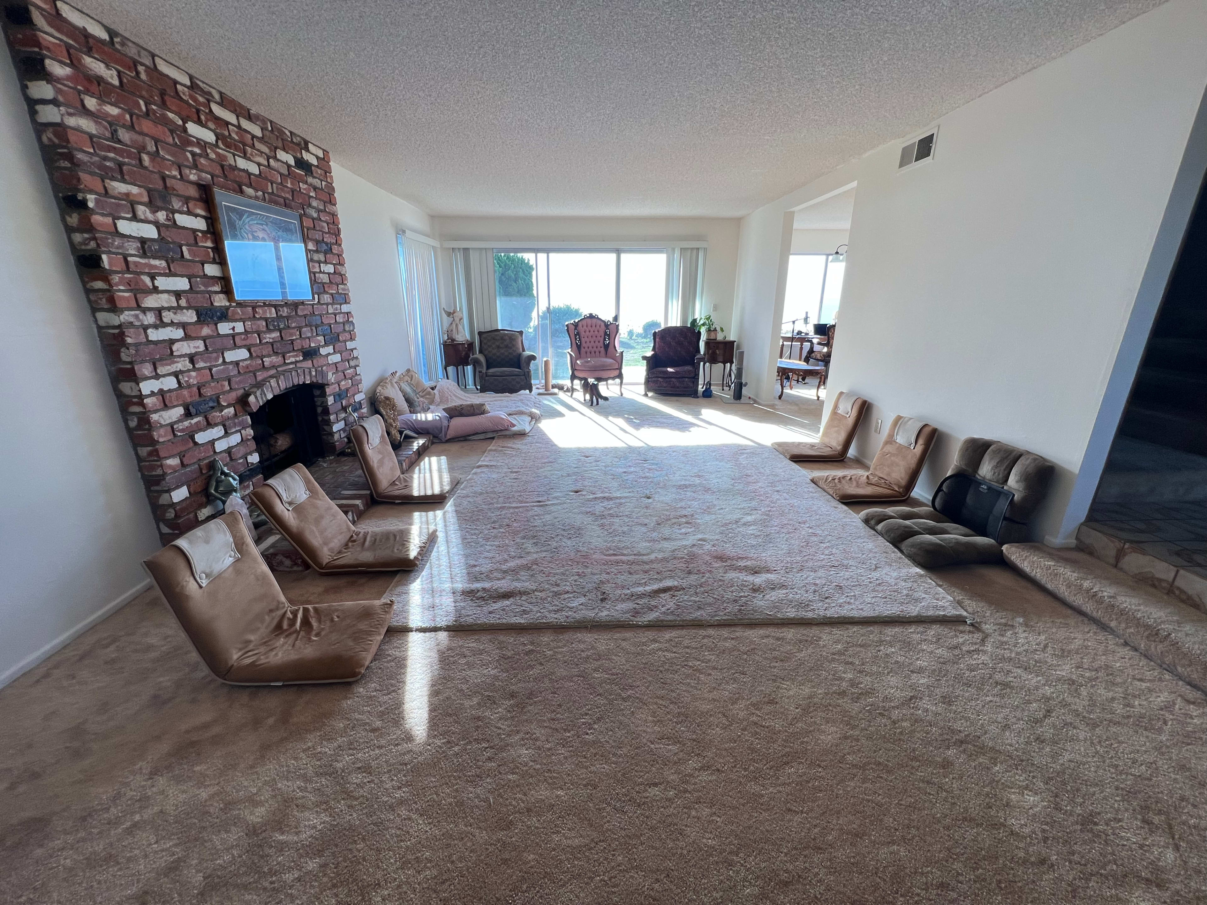The image shows a spacious living area with a large carpet, several floor cushions arranged in front of a brick fireplace, and large windows allowing natural light to fill the room.