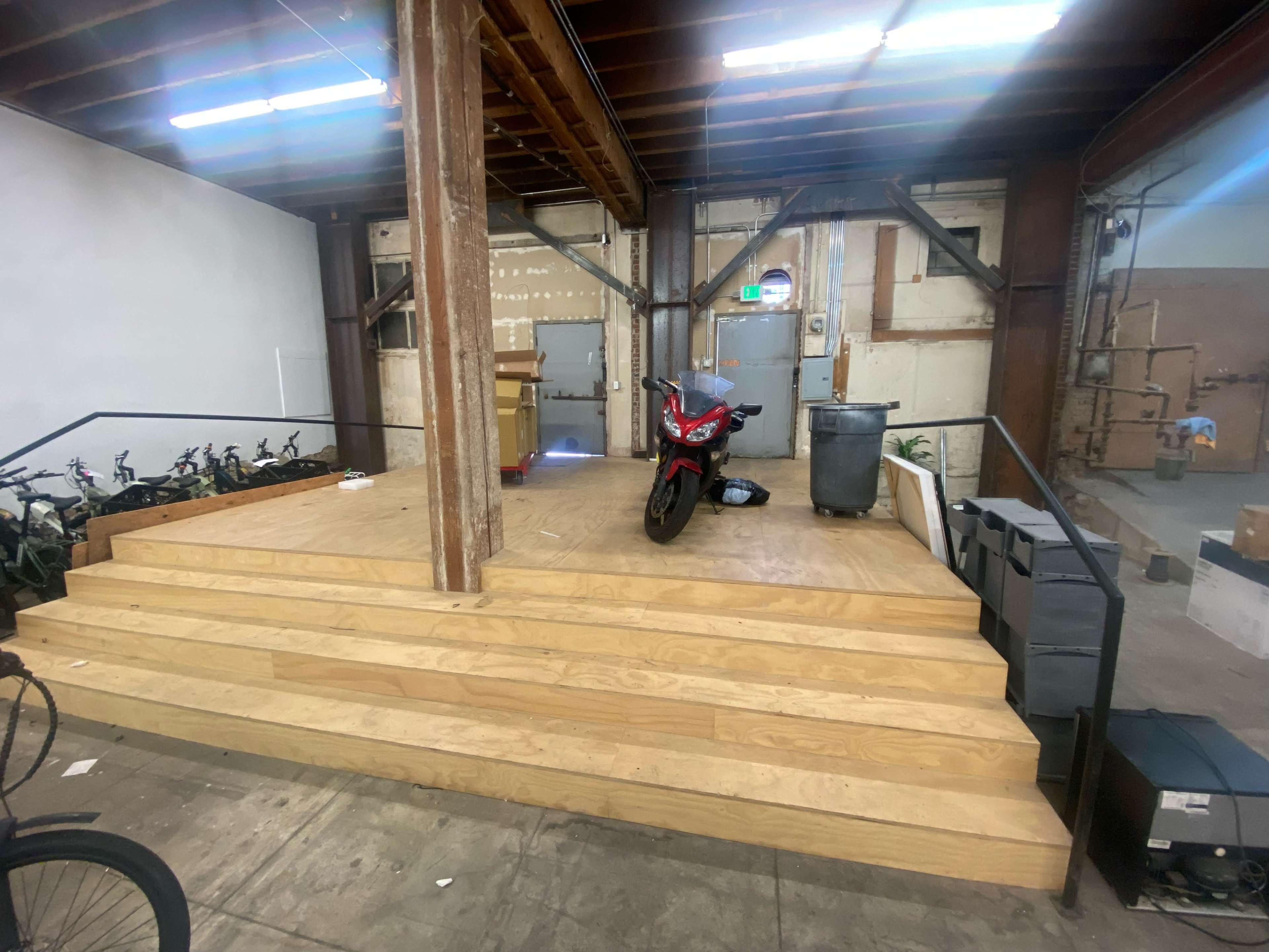 A wooden platform with steps, a motorcycle, and a trash can positioned against a wall in an industrial space.