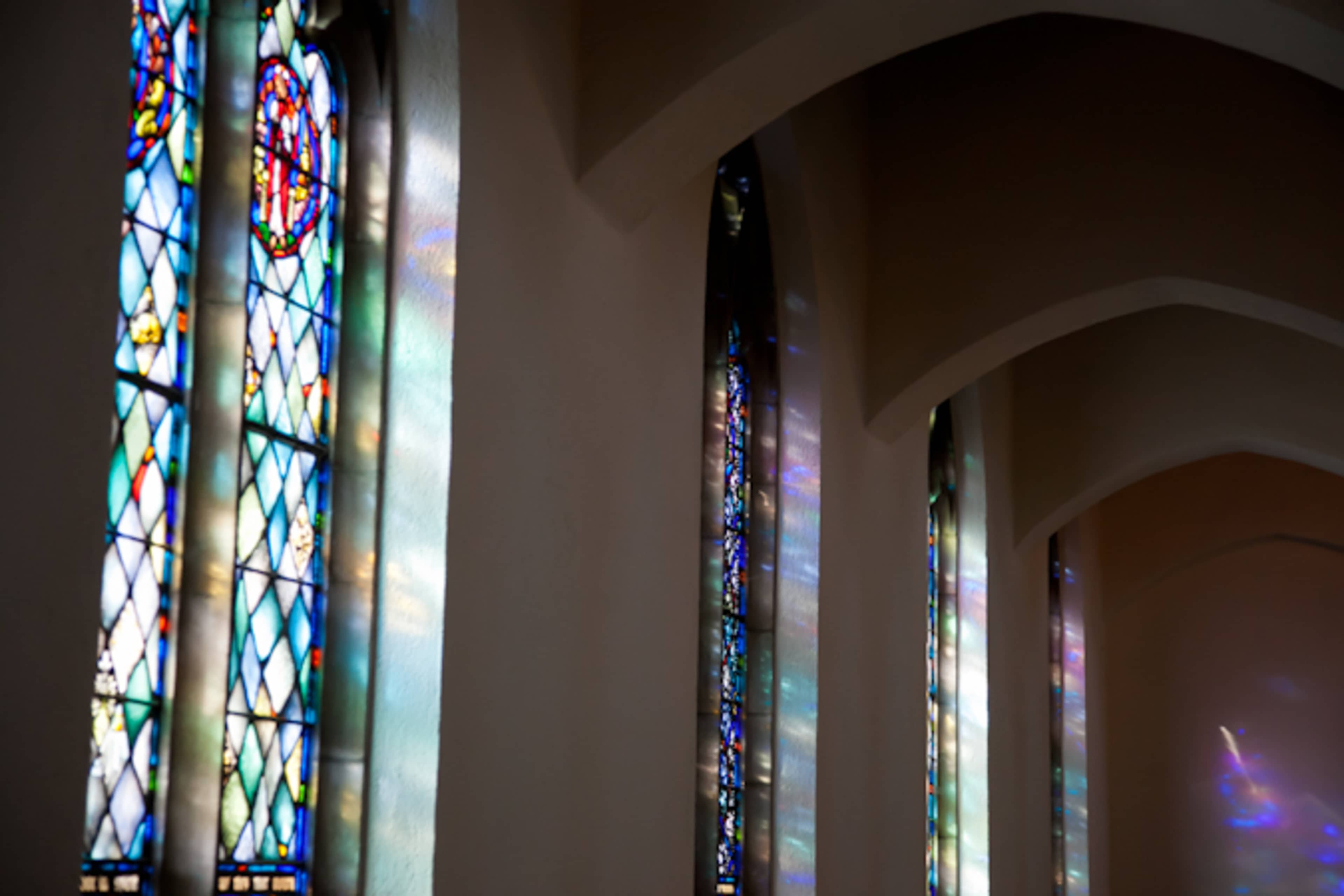 Stained glass windows illuminate a church interior with colorful light patterns on the walls.