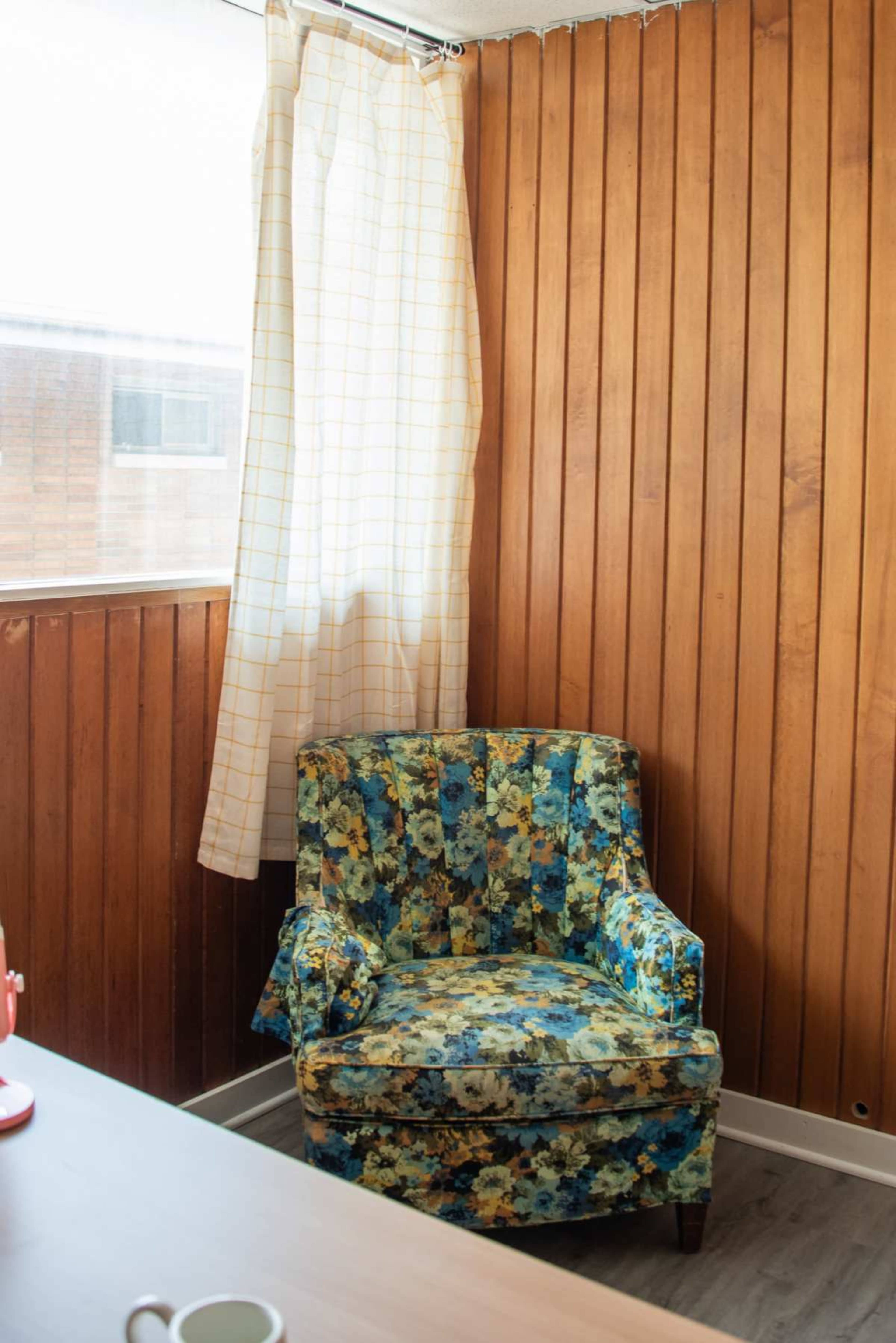 A floral-patterned armchair is positioned in the corner of a room with wood paneling and a sheer curtain.