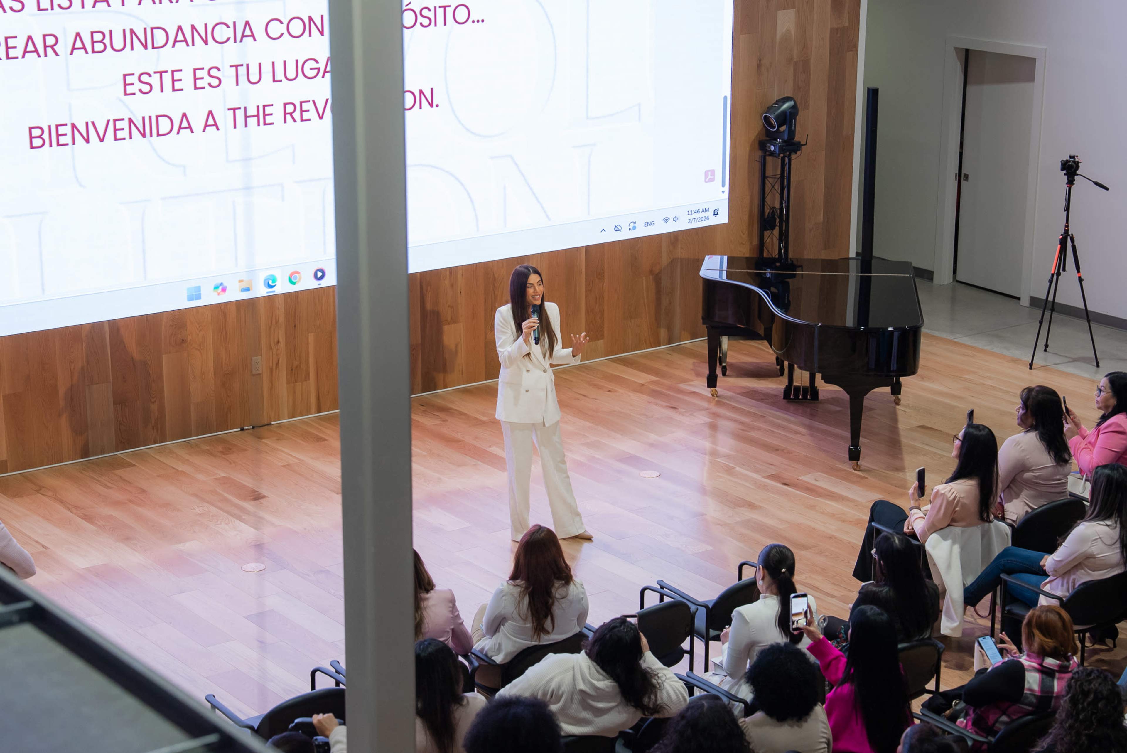 A speaker stands on stage in front of a large screen, addressing an audience seated in rows in a spacious hall with a piano in the background.