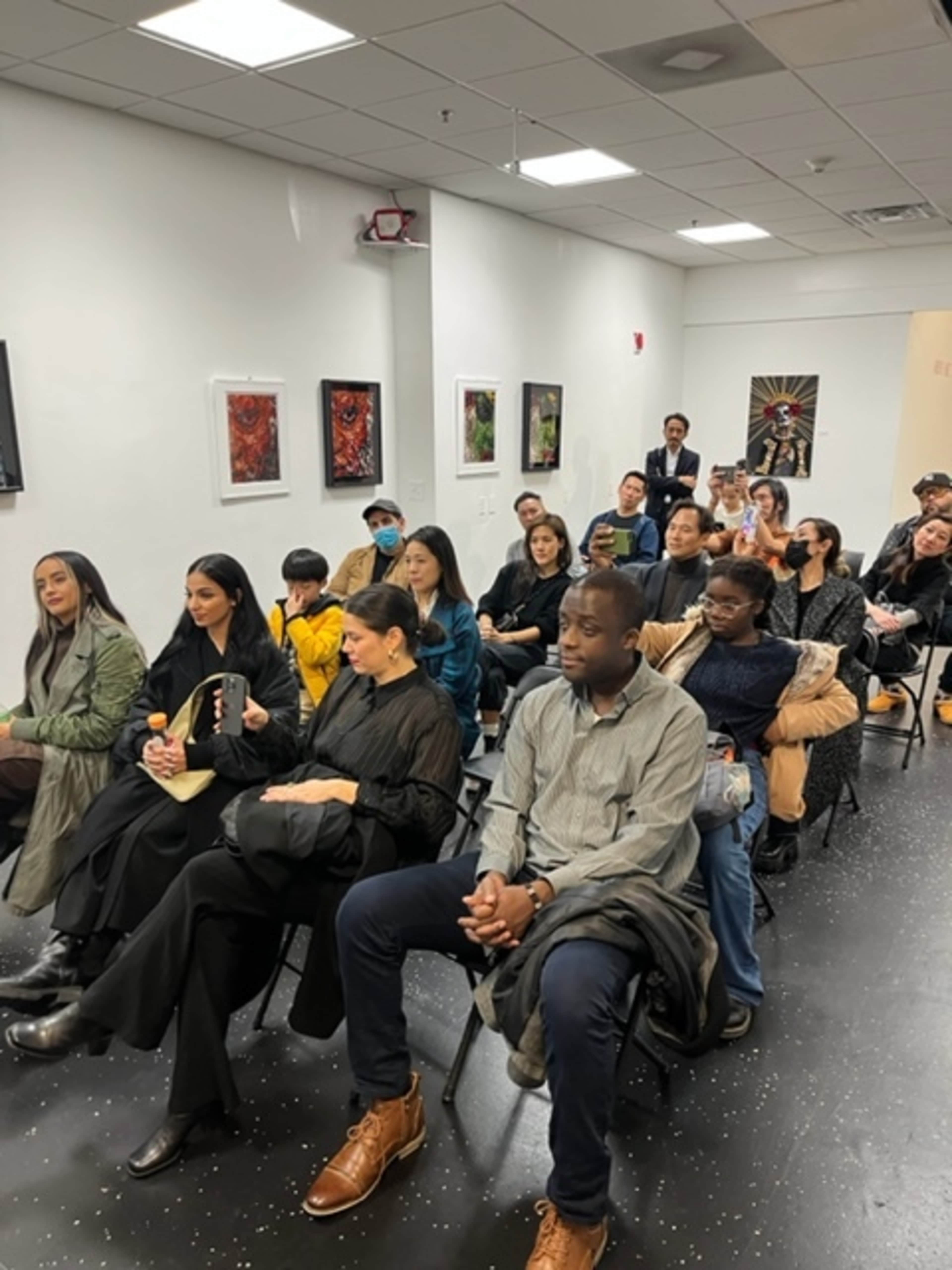 A group of diverse attendees sits in a room, arranged in rows of chairs, facing a front wall adorned with artwork.