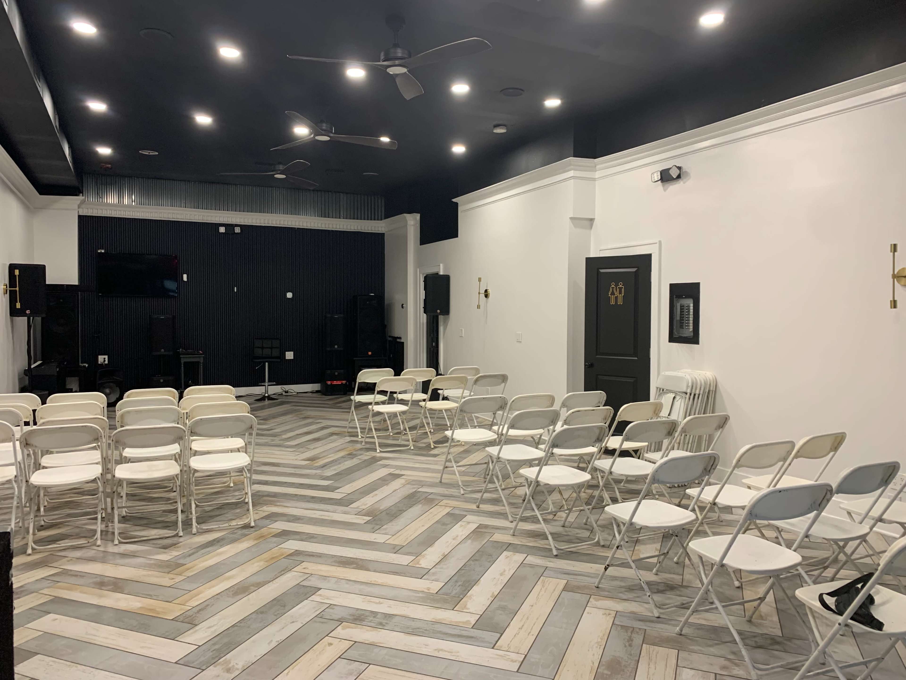 A spacious room with a chevron-patterned floor, set up with white folding chairs facing a small stage and audio equipment on the sides.