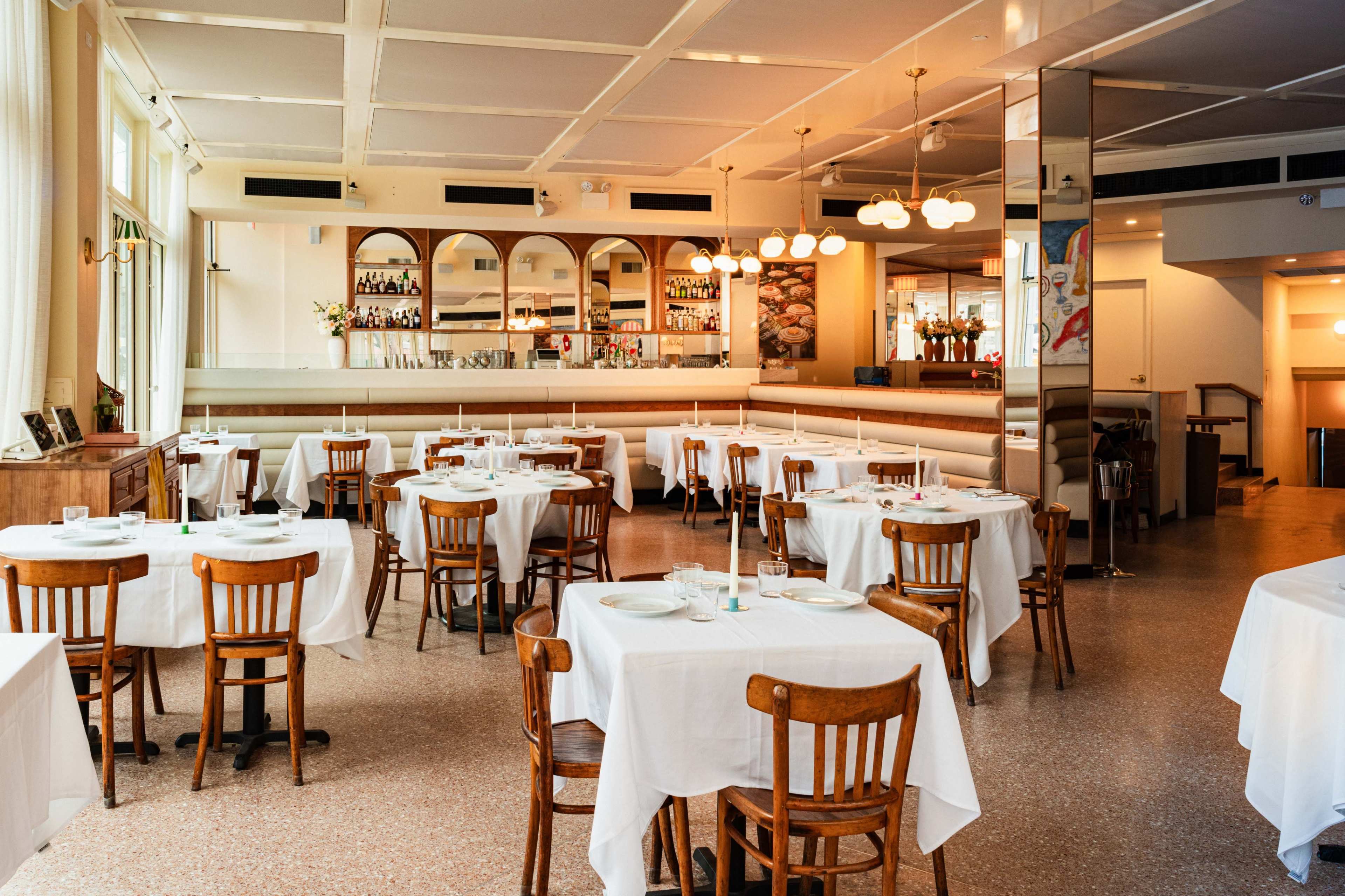 The image shows a spacious restaurant with wooden chairs and white tablecloths arranged around neatly set tables, illuminated by soft lighting and featuring a bar area in the background.