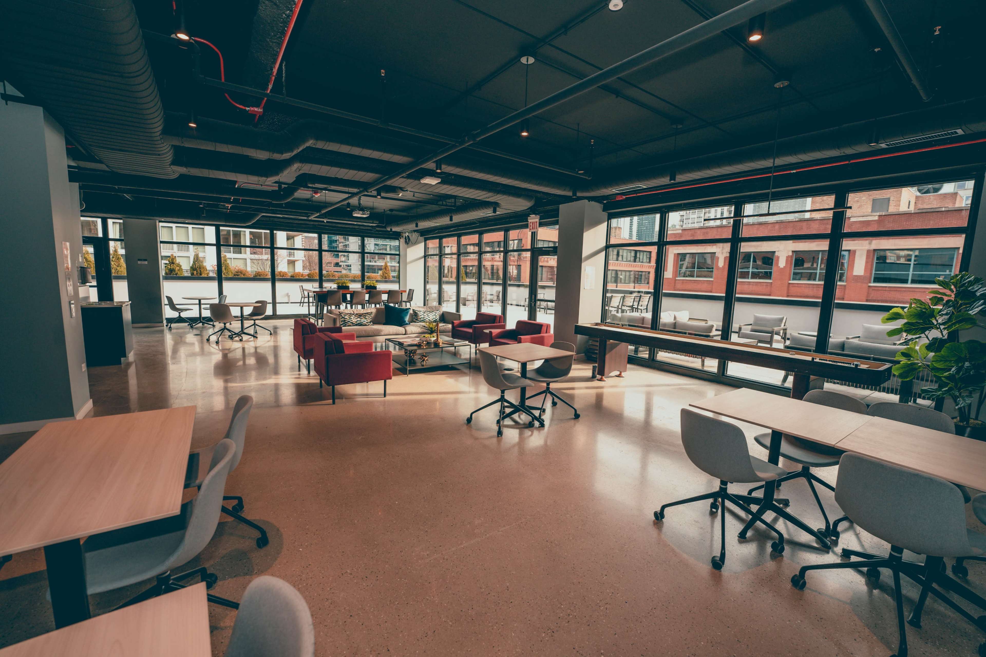 A modern office space with large windows, multiple seating areas, and various tables arranged throughout.