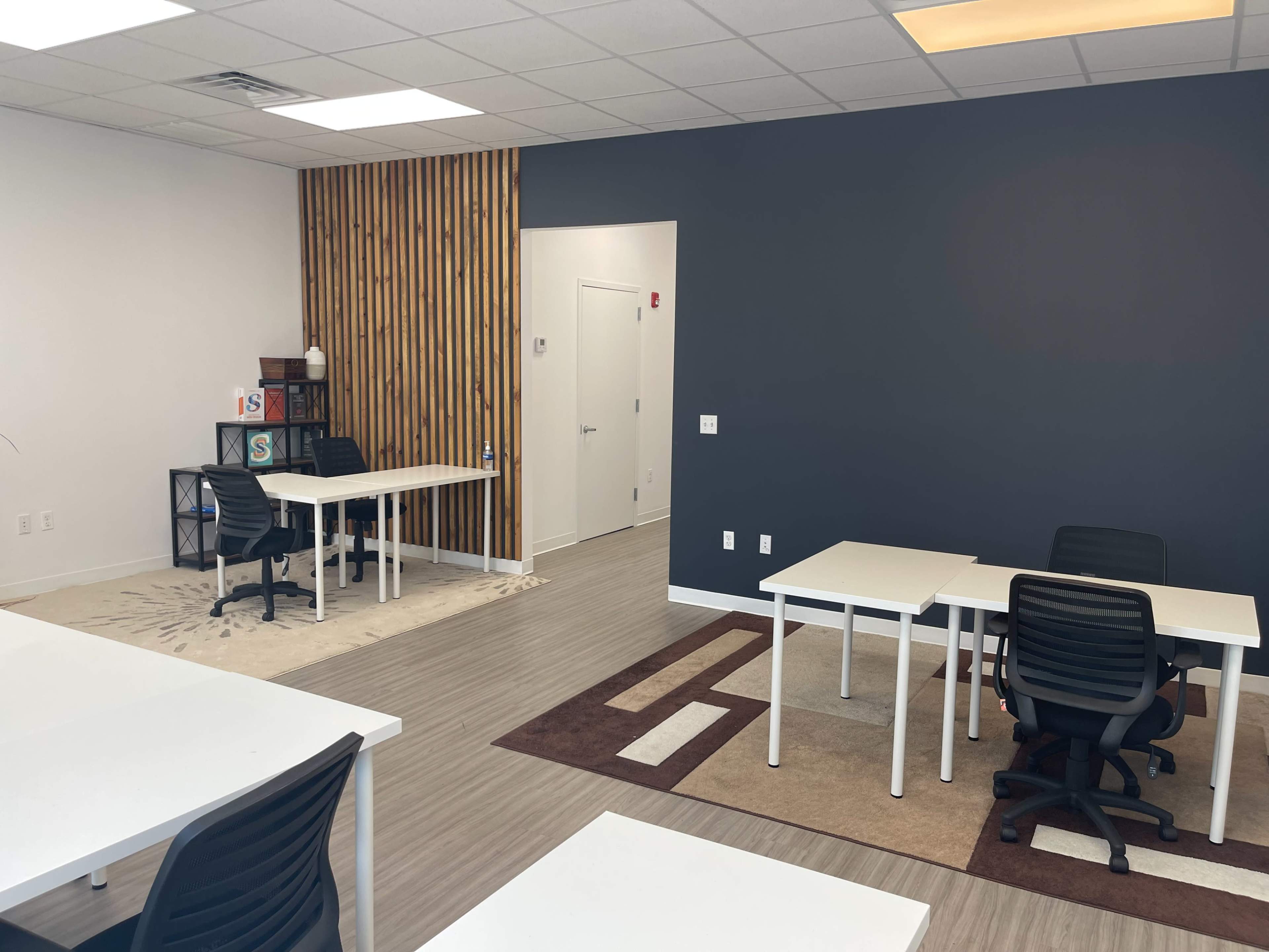 The image shows an office space with several white desks and black chairs, set against a backdrop of a dark gray wall and a wooden accent wall.