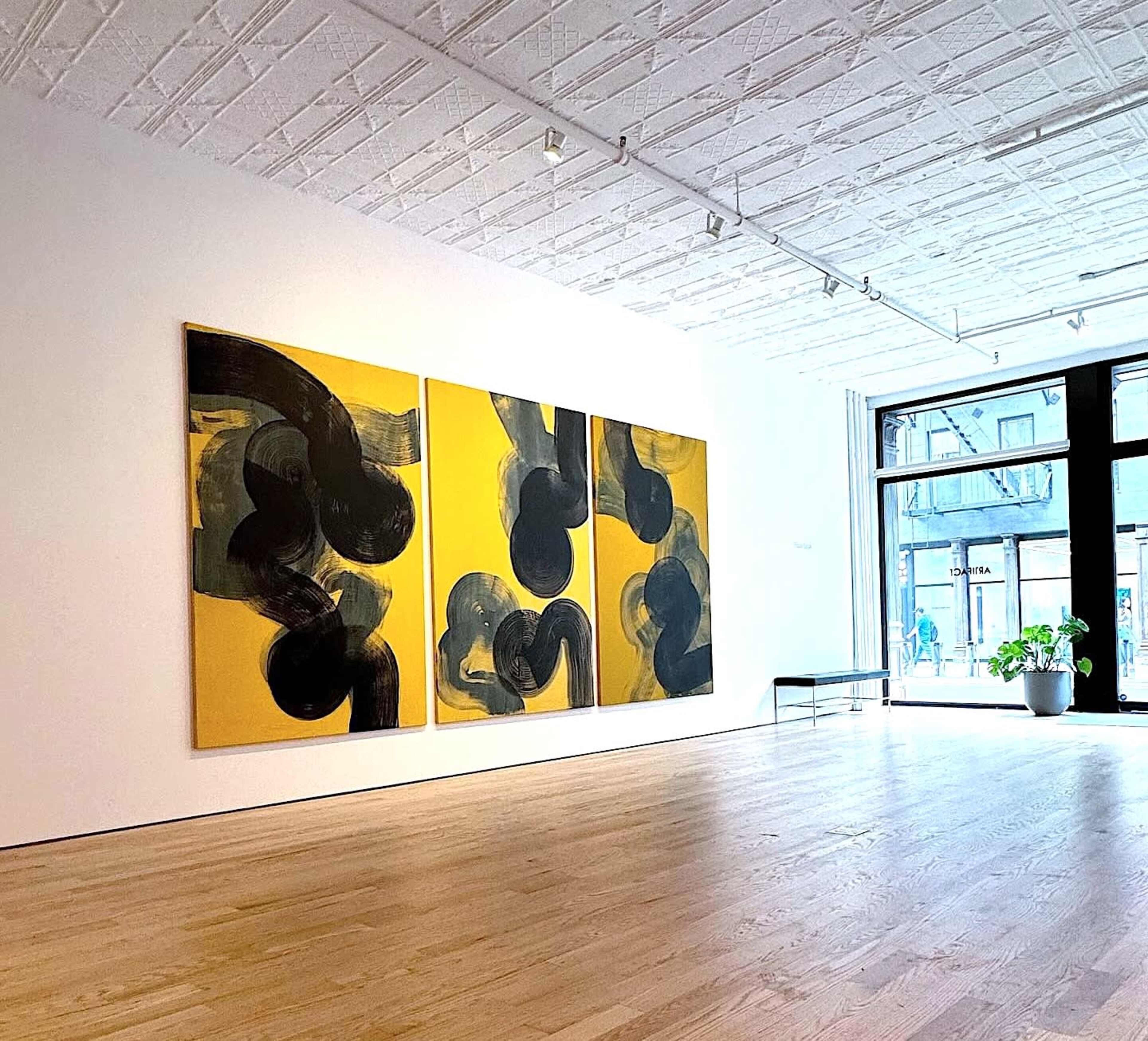 A spacious art gallery displays a large abstract triptych with bold black shapes on a yellow background, complemented by wooden flooring and large windows.