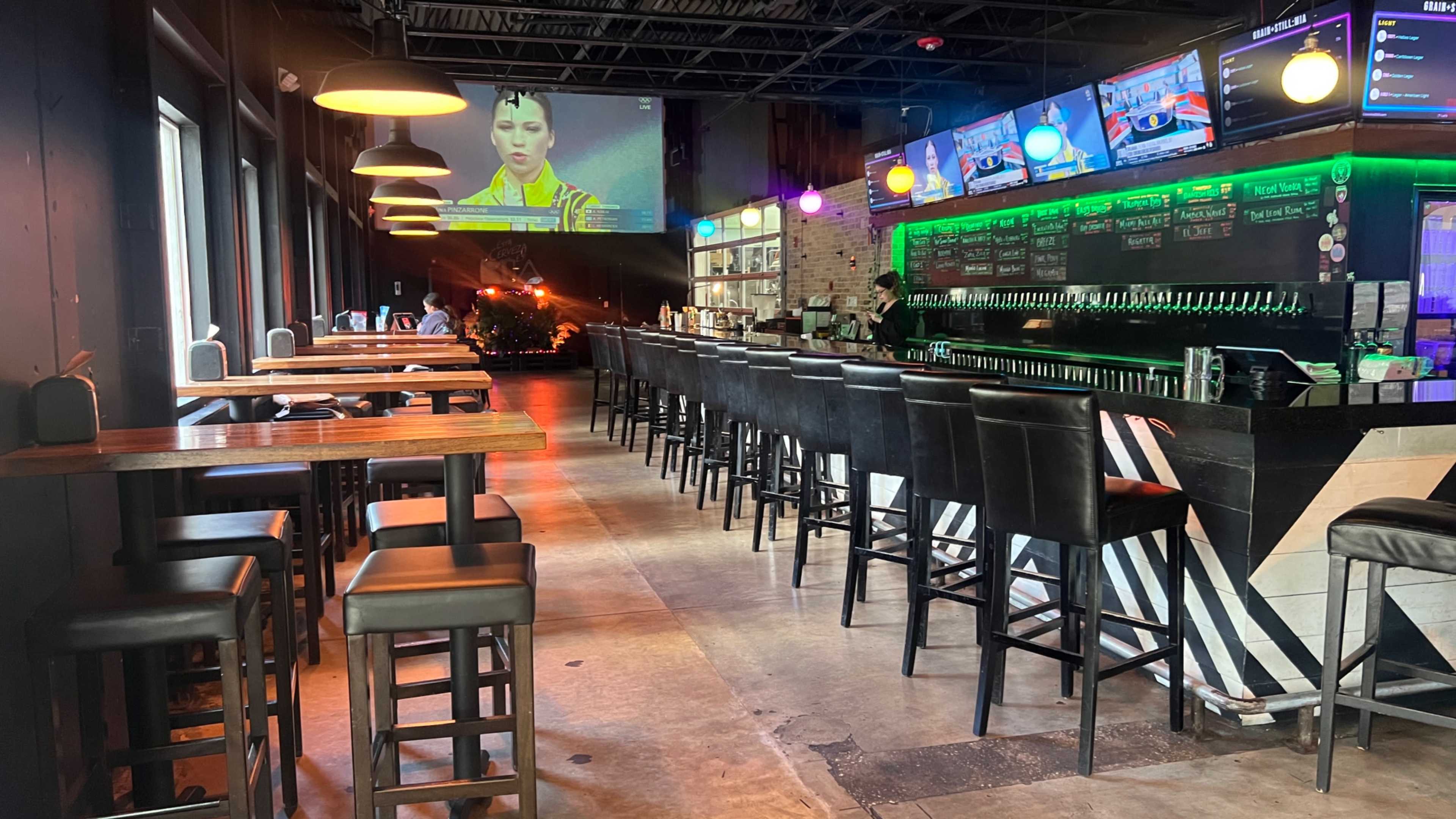A well-lit bar with high stools and multiple screens displaying a video, accompanied by a row of beer taps along the back wall.