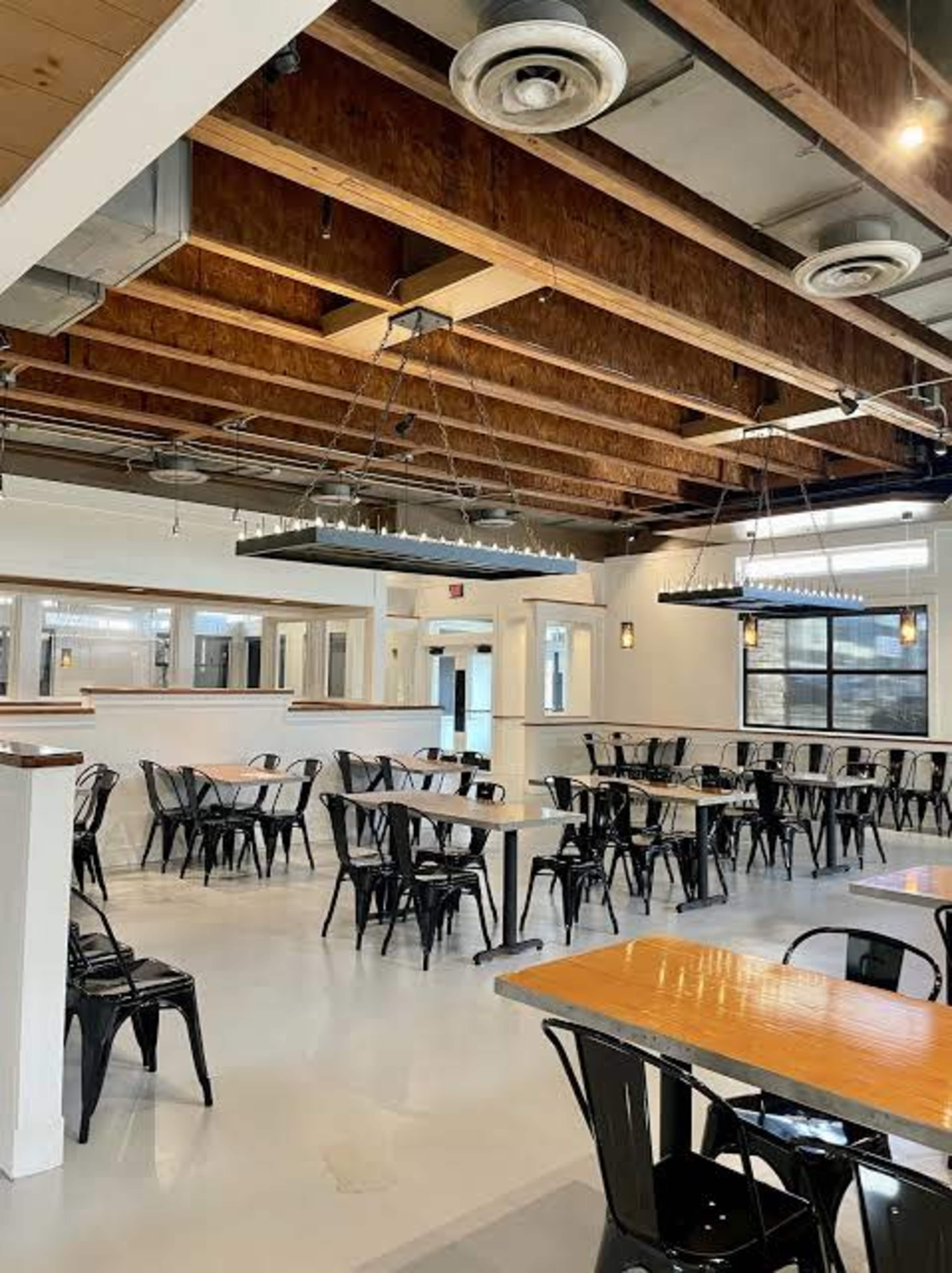 The image shows a spacious dining area with metal chairs and wooden tables, featuring exposed beams in the ceiling and large windows allowing natural light.