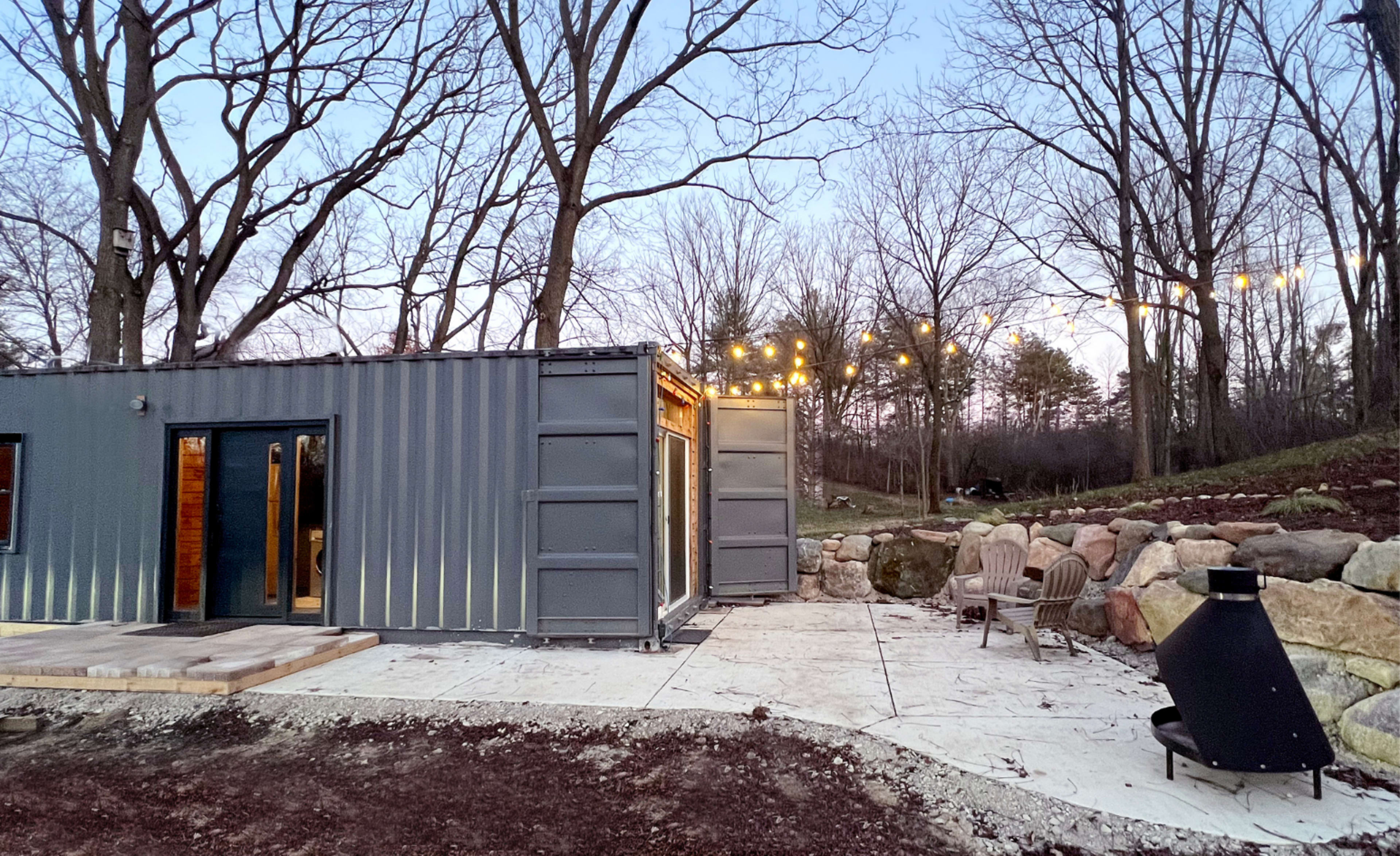 A modern container home surrounded by trees, with patio lights strung overhead and a fire pit along a stone wall.