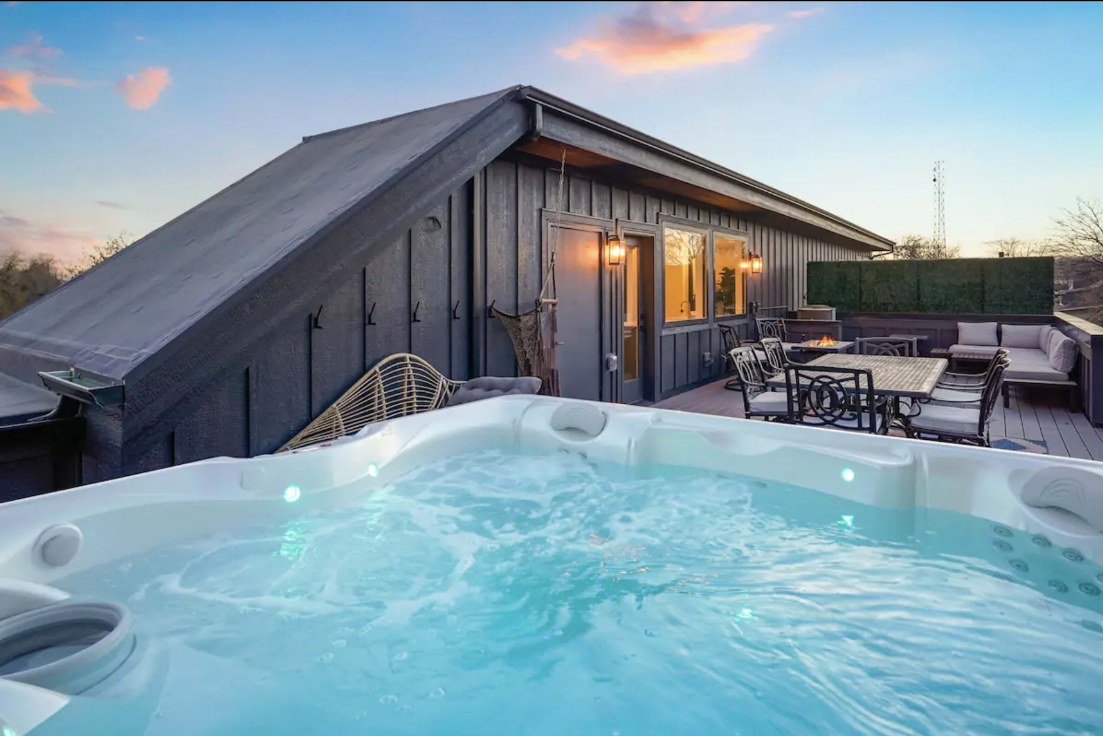 A hot tub filled with water sits on a wooden deck next to a modern building under a colorful sky at sunset.