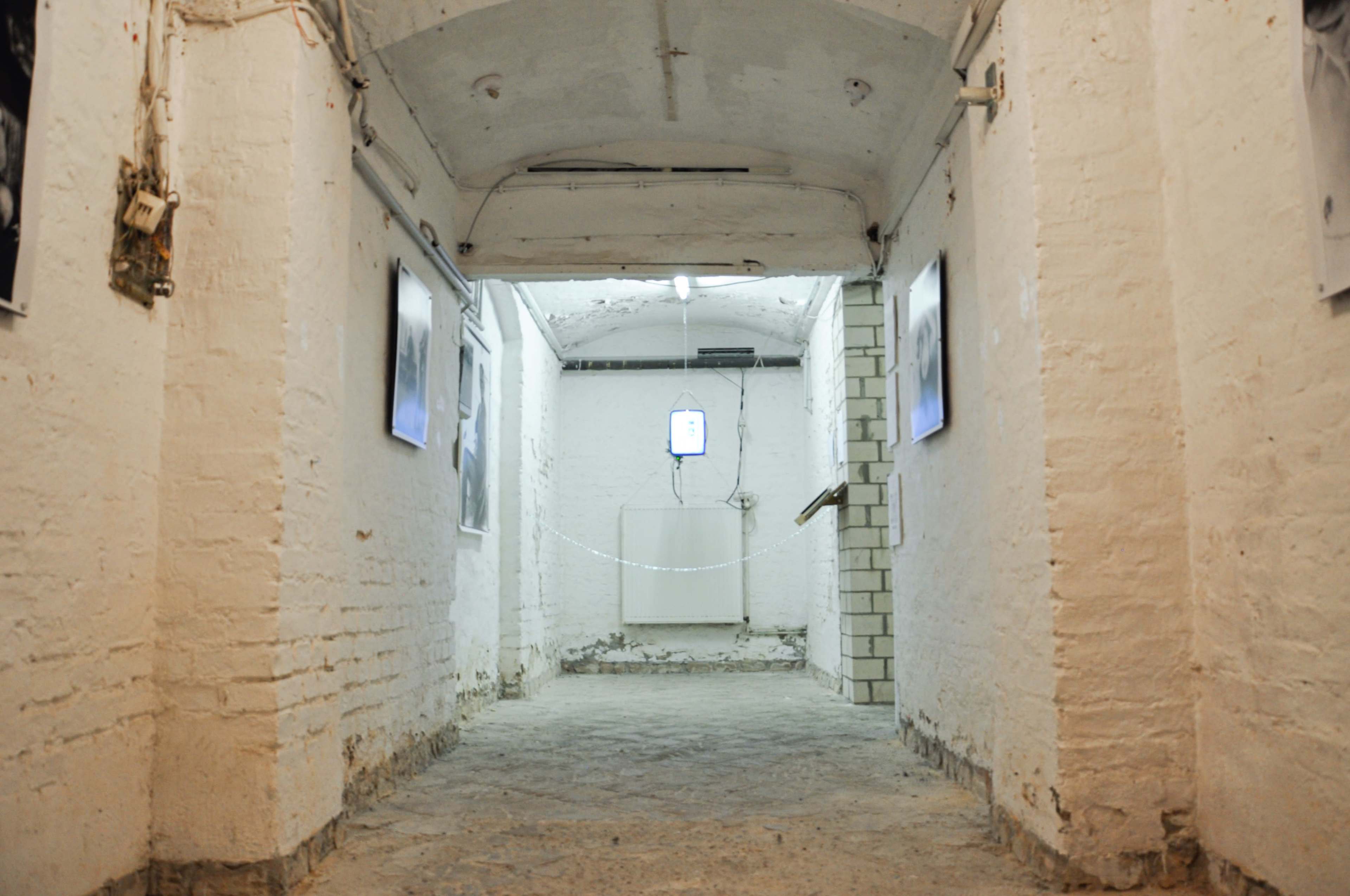 A narrow, whitewashed corridor with exposed brick walls features illuminated screens on either side and a door at the far end.