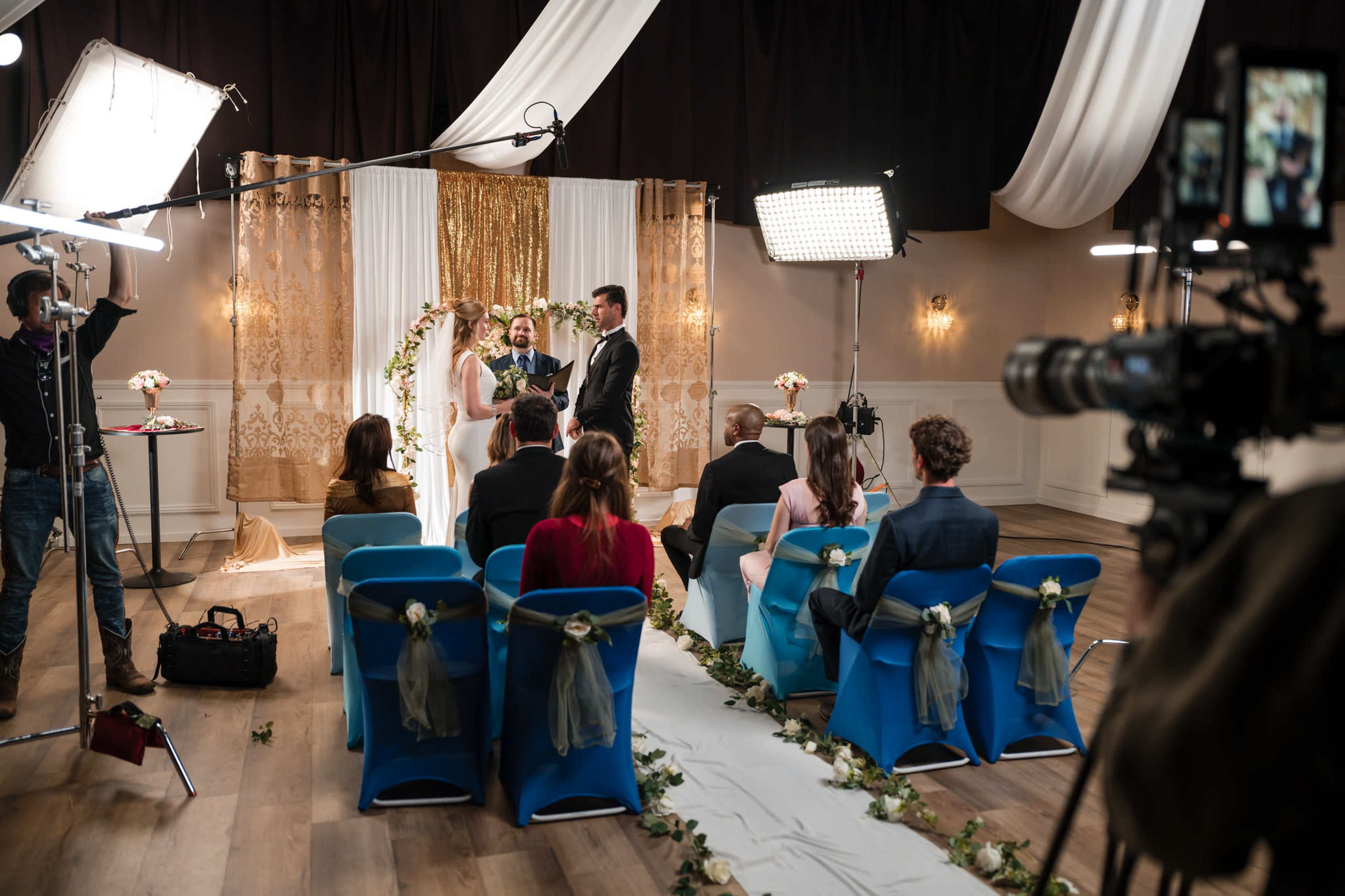 A couple is exchanging vows during a wedding ceremony in a decorated venue, while guests sit in blue chairs.