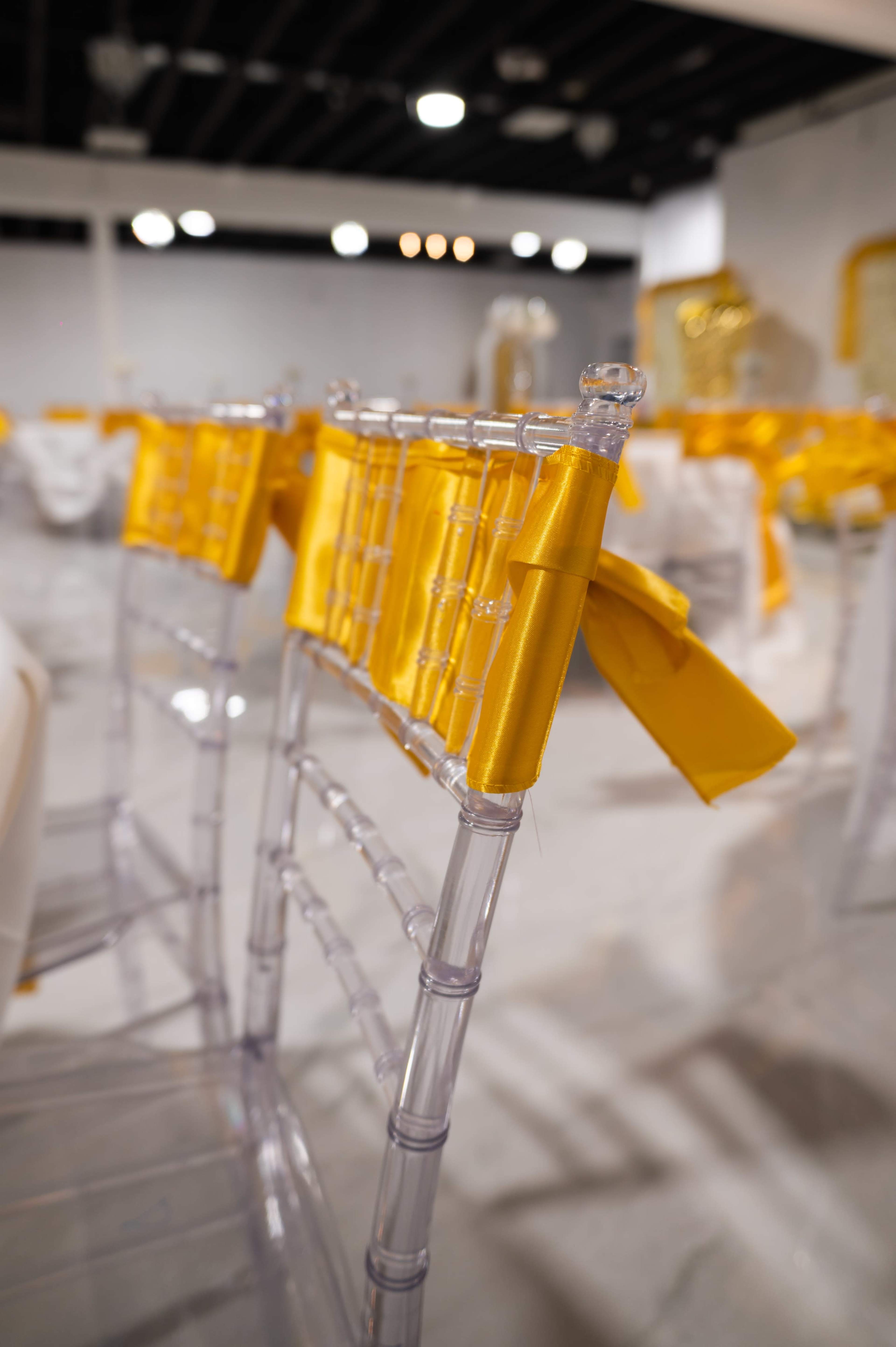 The image shows clear chairs with yellow ribbons tied to the backs, set in a decoratively arranged indoor event space.