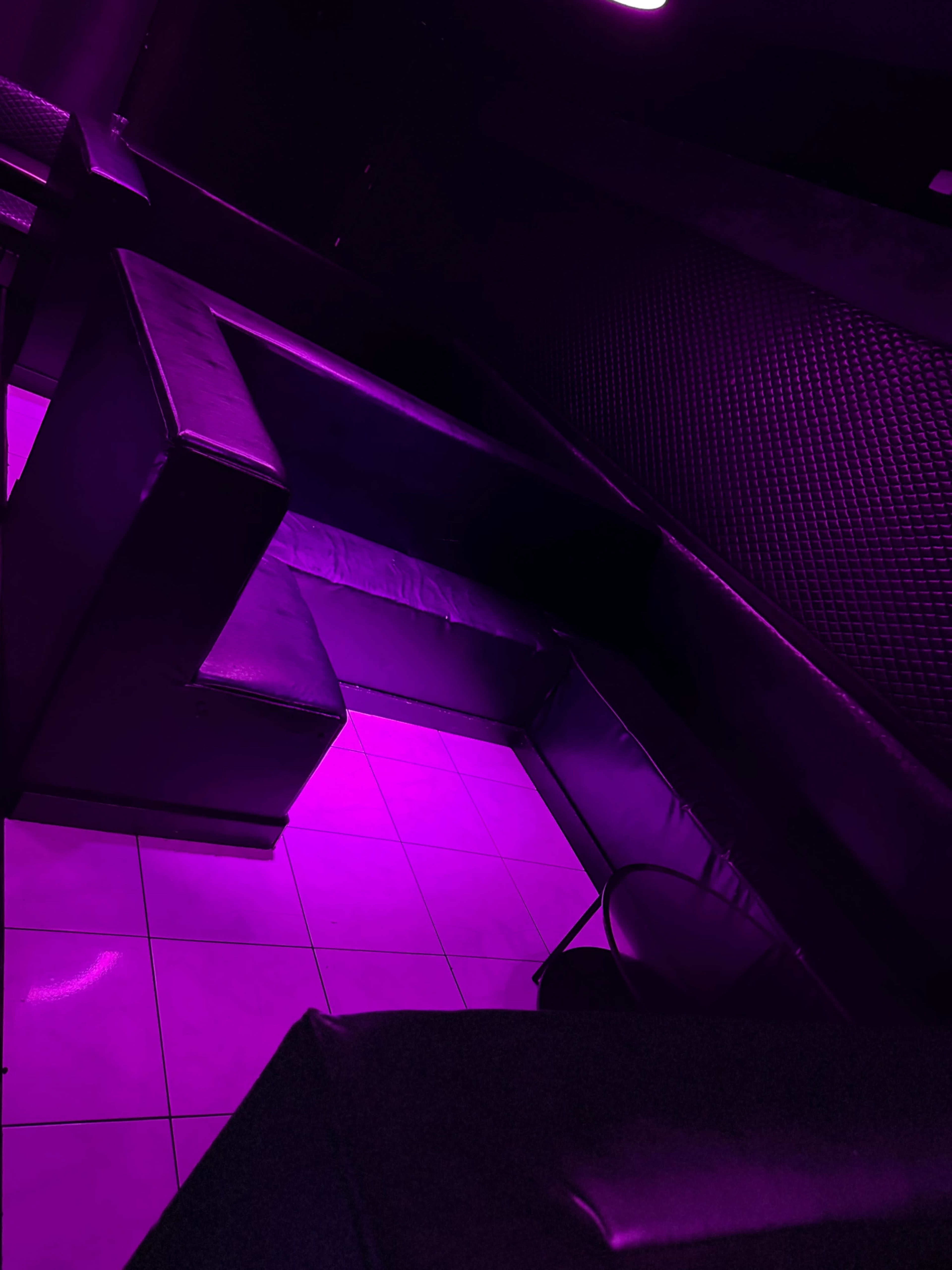 The image shows a dimly lit lounge area with purple lighting, featuring black leather seating arranged along the walls and a tiled floor.