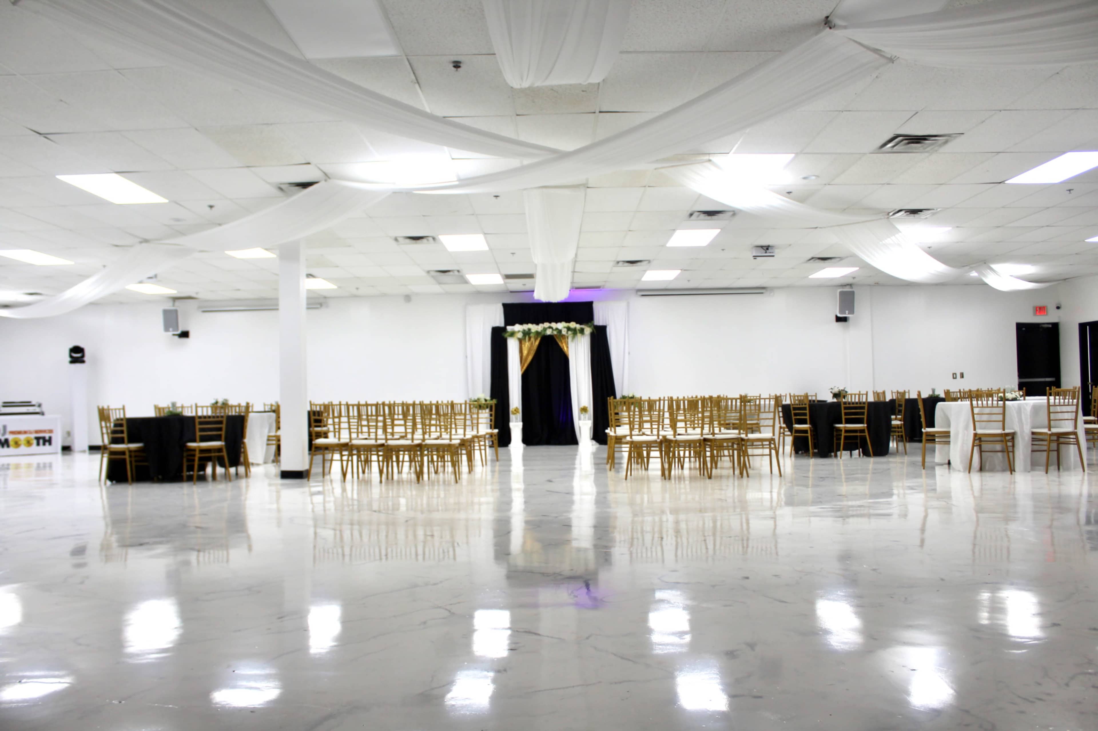 A spacious event hall features rows of gold chairs, tables set for dining, and decorative drapery above.