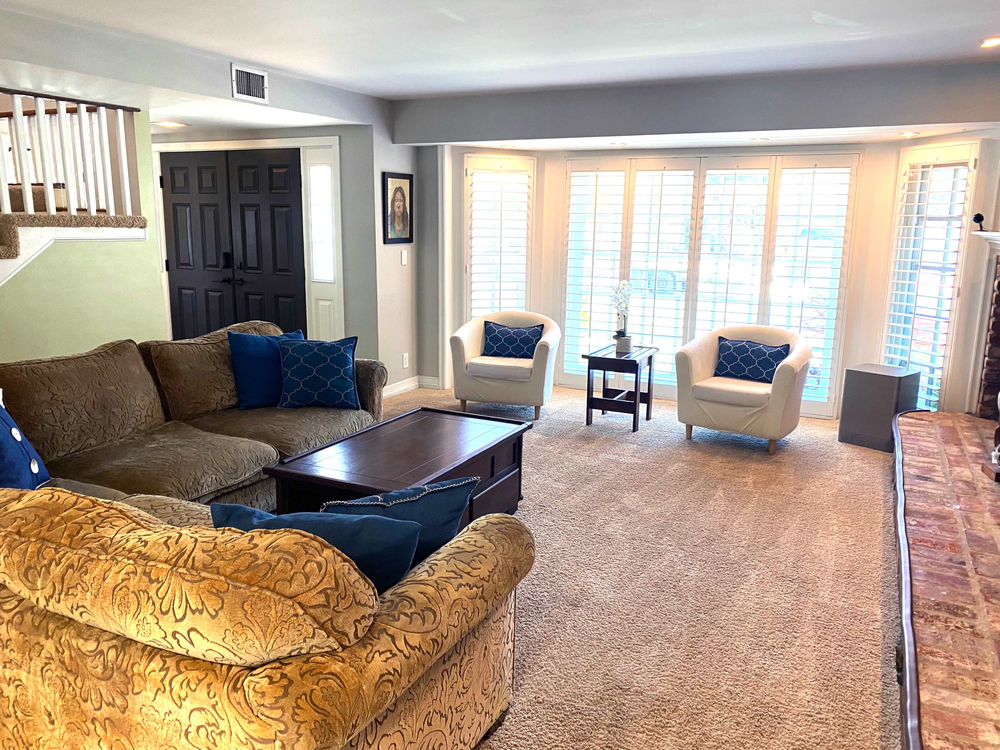 The living room features a large, patterned couch, two white armchairs, a dark coffee table, and bright windows with plantation shutters.