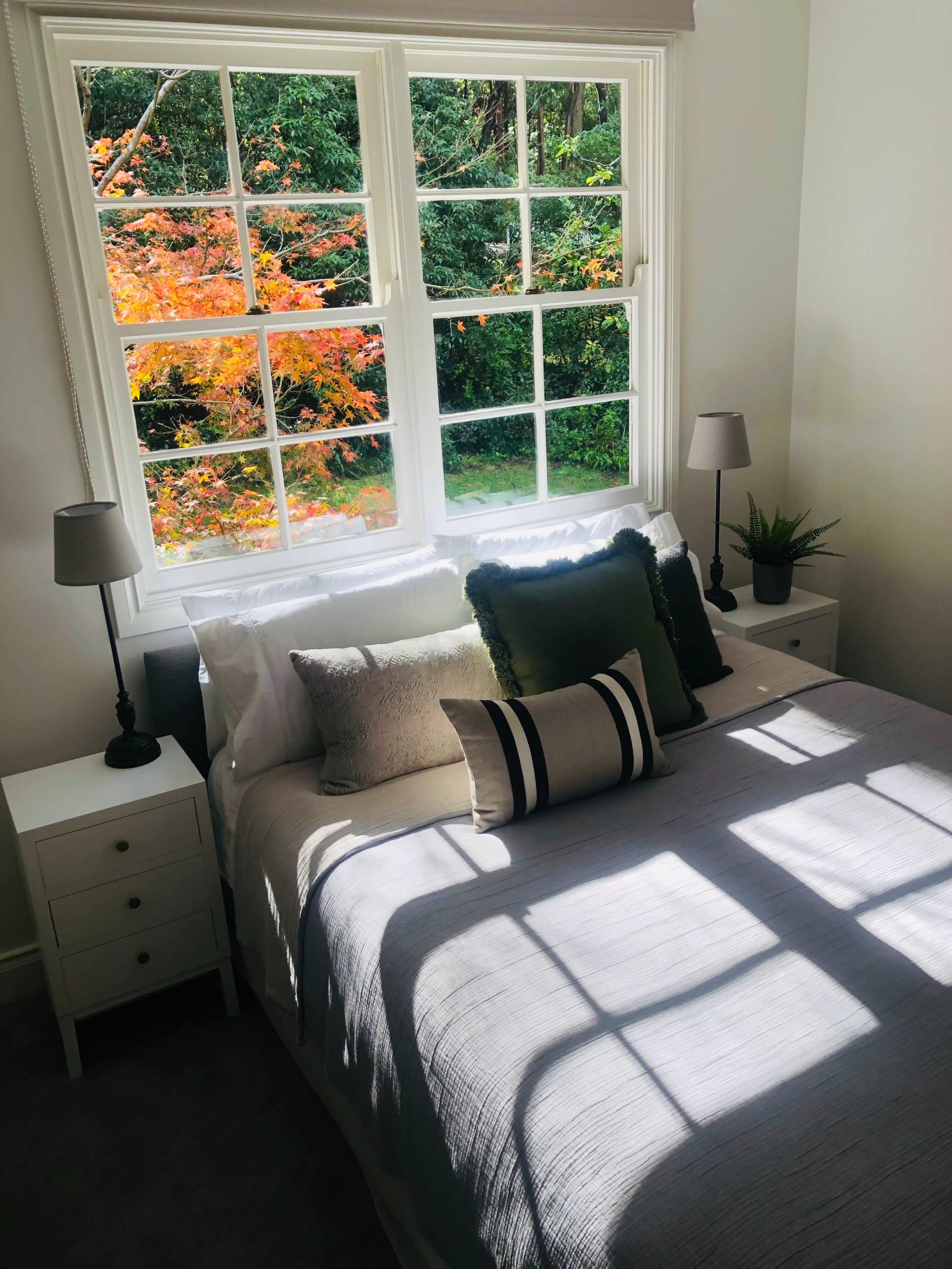 A well-lit bedroom features a bed with decorative pillows, bedside tables with lamps, and a view of colorful foliage through the window.