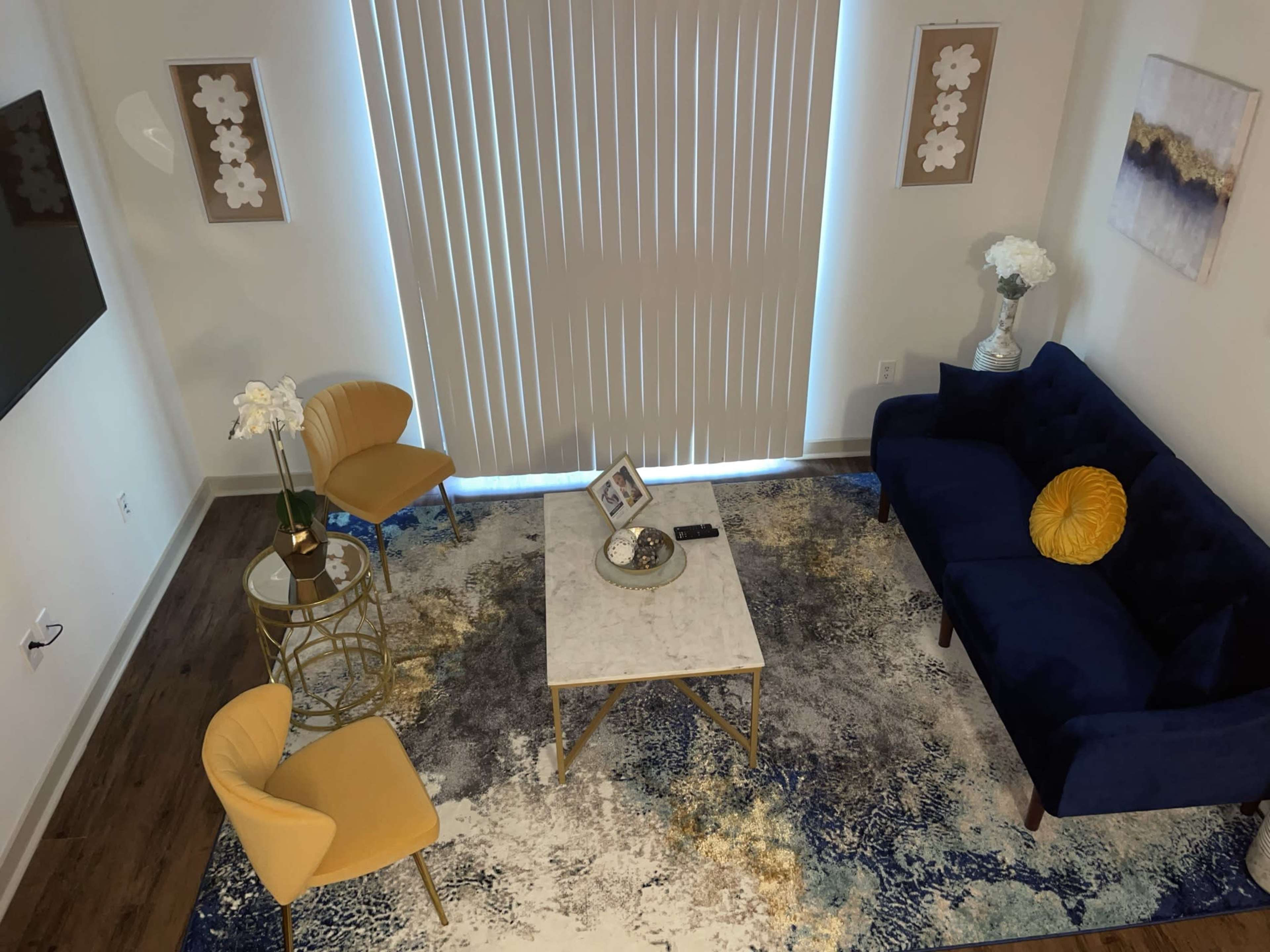 A cozy living room setup with a blue sofa, two yellow chairs, a marble coffee table, and decorative elements on a blue and white rug under vertical blinds.